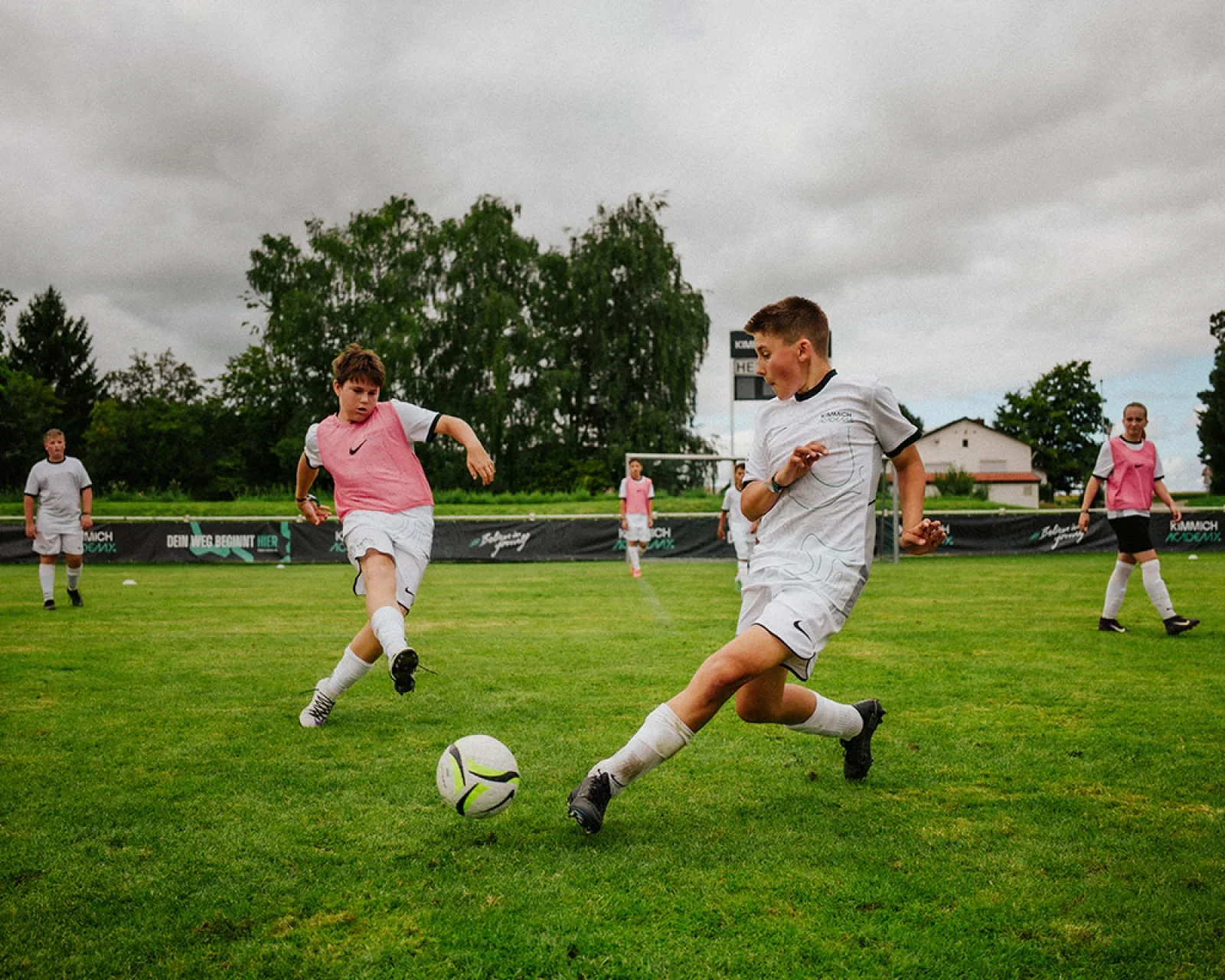 Young soccer players practicing on an outdoor field under cloudy skies, with a boy kicking a ball in the foreground and others watching or waiting in the background.