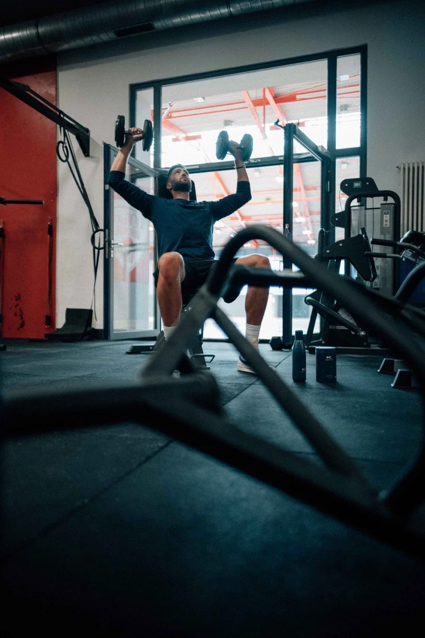 A man lifting dumbbells in a gym with exercise equipment and a large window behind him.