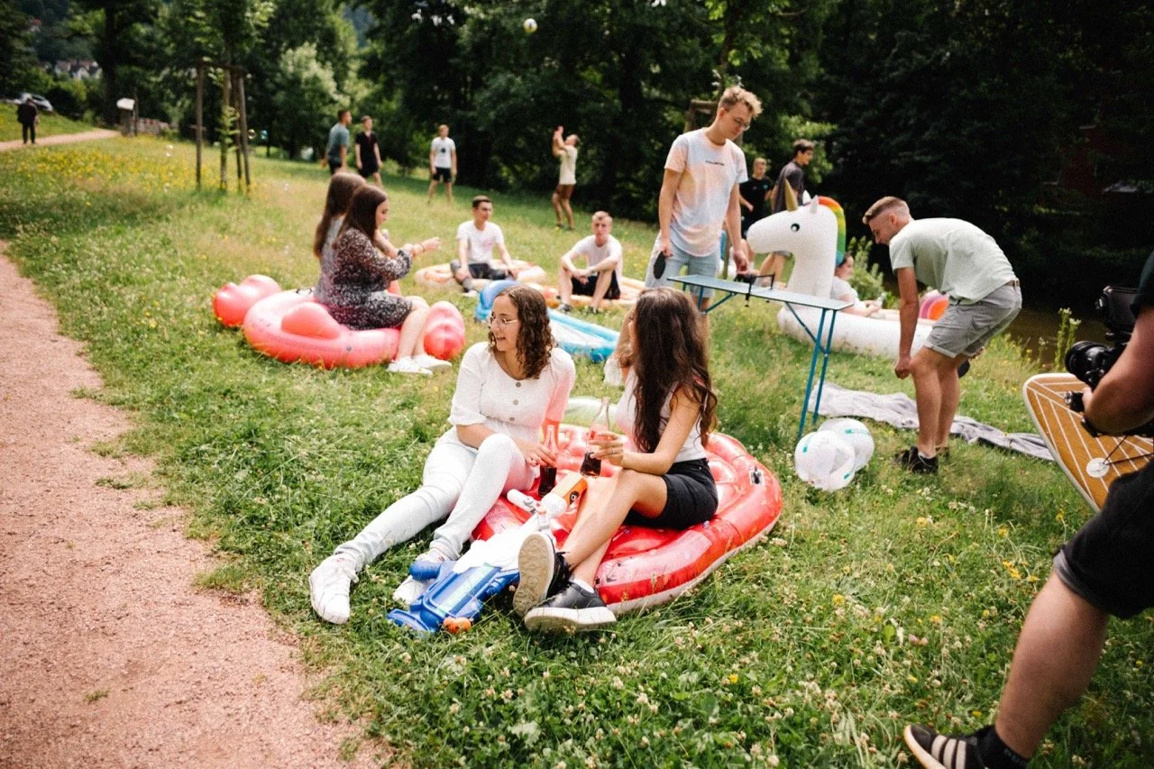 Group of young people having a picnic and relaxing on inflatable floats by a river in a park on a sunny day, with some people standing and others sitting on grass or inflatables.