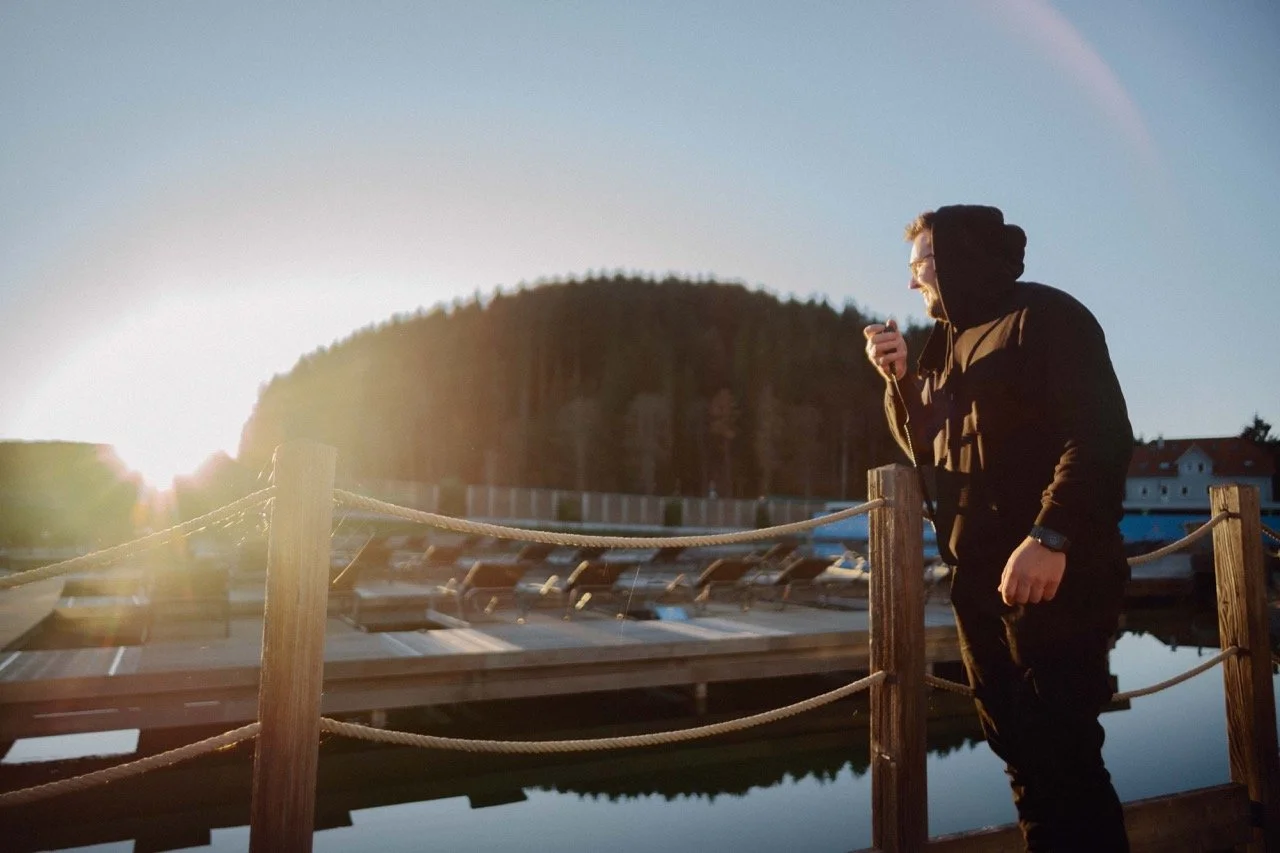 A man wearing a black hoodie standing on a dock at sunset, looking at his phone, near boats and a body of water, with trees and buildings in the background.