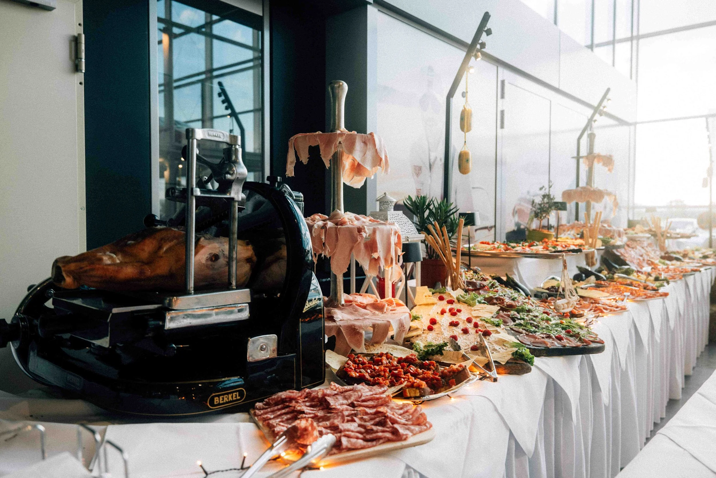 Buffet table with various dishes including meats, cheeses, and vegetables, set in a bright room with large windows.