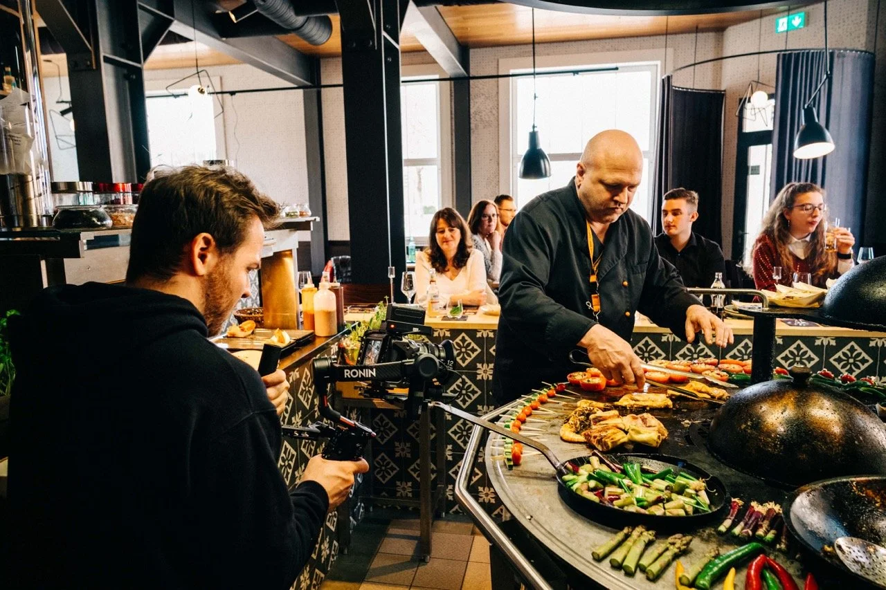 Chef cooking grilled vegetables and chicken at a restaurant kitchen, with a cameraman recording the process and diners seated in the background.