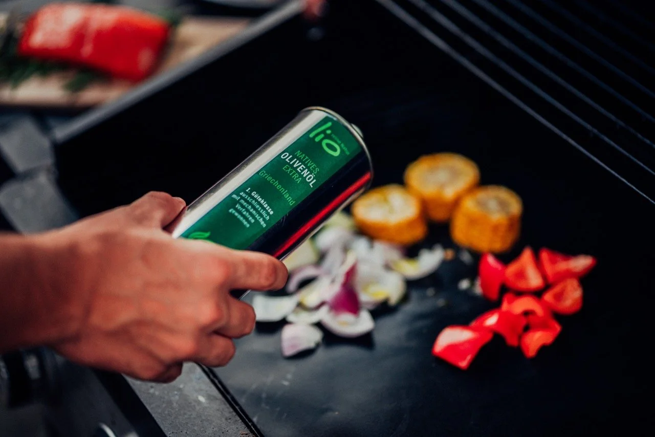 Person pouring olive oil from a green bottle onto vegetables grilling on a barbecue, including sliced zucchini, red bell peppers, and onions.