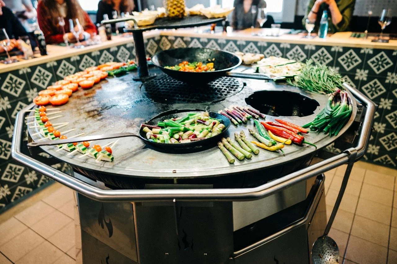 Tabletop with assorted grilled vegetables and skewers including tomatoes, peppers, artichokes, and green beans, with diners in the background.