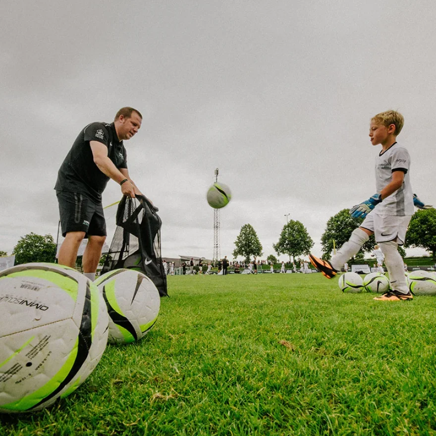 A man and a young boy, likely a coach and a player, are standing on a soccer field with multiple soccer balls around them. The man is holding a sports bag, and the boy, wearing a white kit and gloves, is preparing to kick a soccer ball. The scene is 