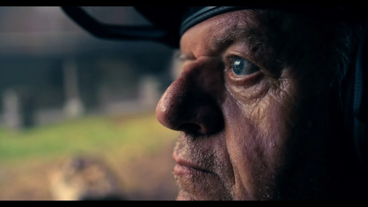 Close-up side profile of an elderly man with a rugged face, wearing a dark cap, looking out through a vehicle window.