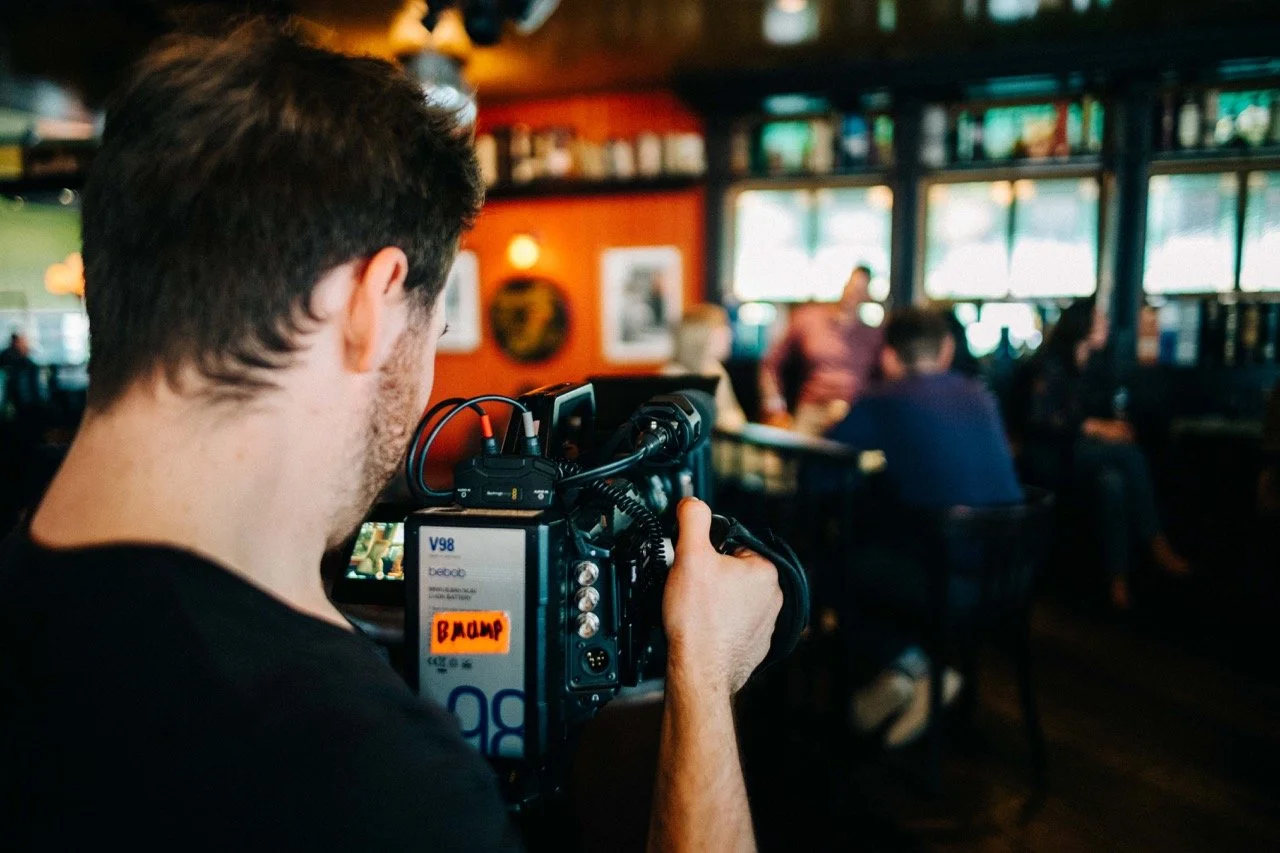 A man filming a conversation at a table in a busy restaurant or bar with orange walls, framed pictures, and windows in the background.