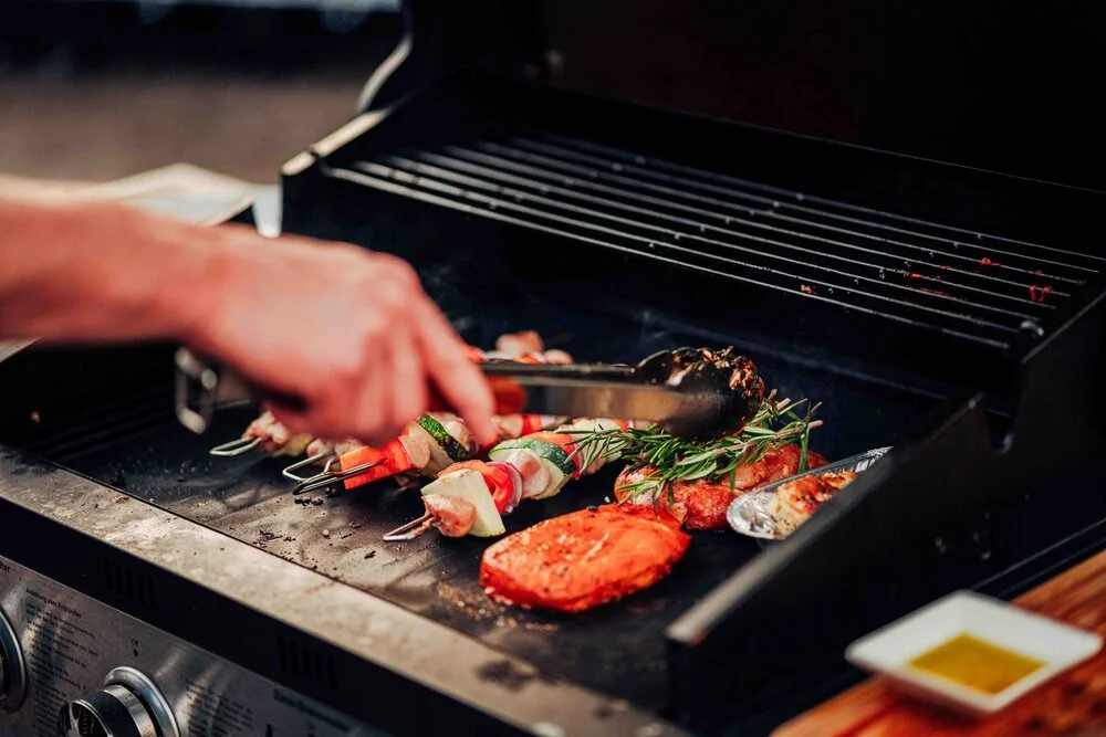 Person grilling skewers of meat and vegetables with rosemary on a barbecue grill.