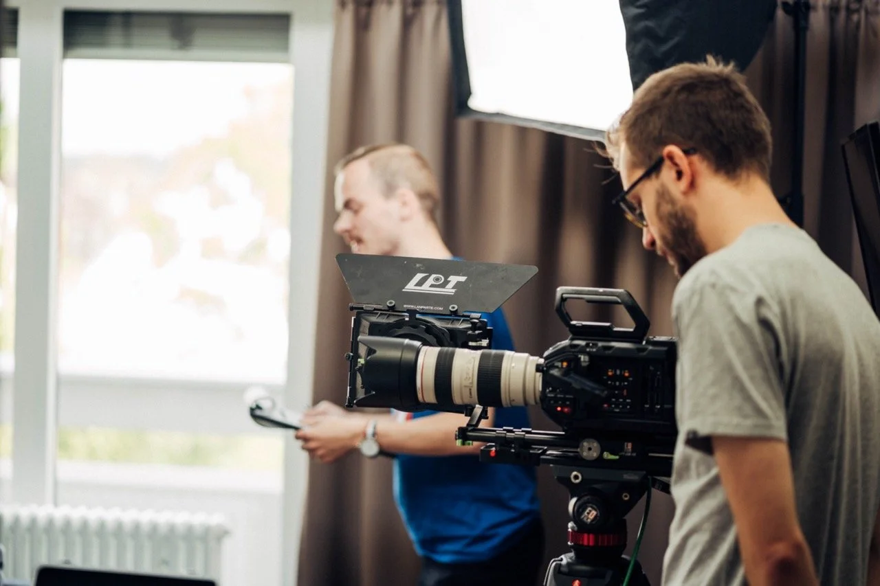 Two men working with video production equipment indoors, with large window and curtains in the background.
