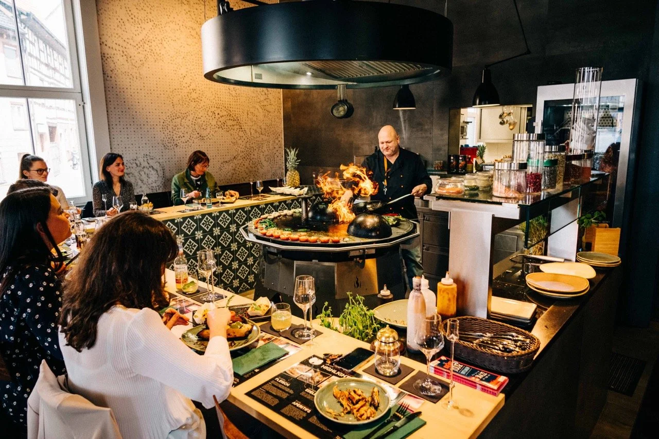 Chef preparing food on a flaming grill station while guests sit at a long table in a modern restaurant.
