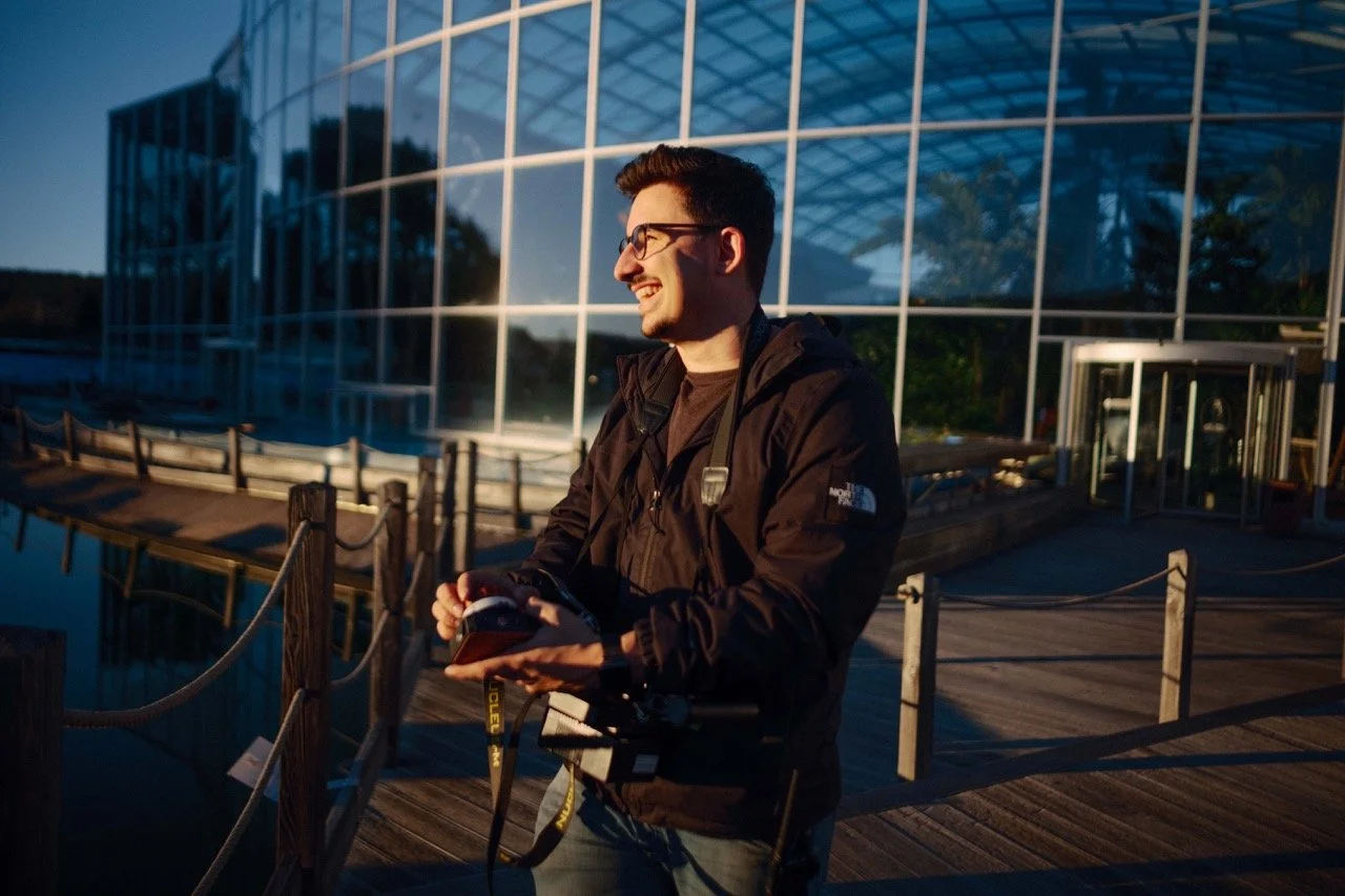 A young man smiling and holding a drone controller, standing on a wooden dock outside a modern glass building in the evening.