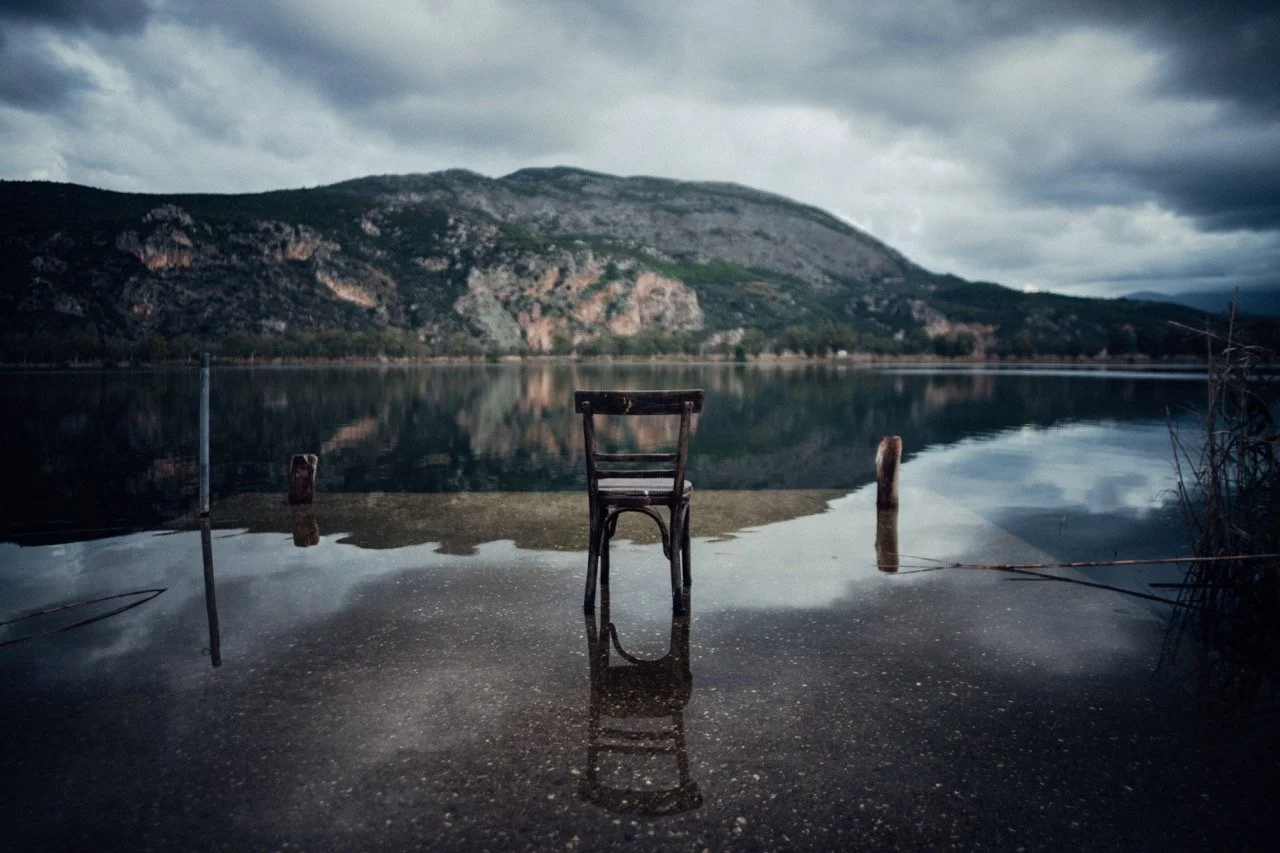 A solitary wooden chair partially submerged in water on a lakeshore, with mountains in the background and dark cloudy sky