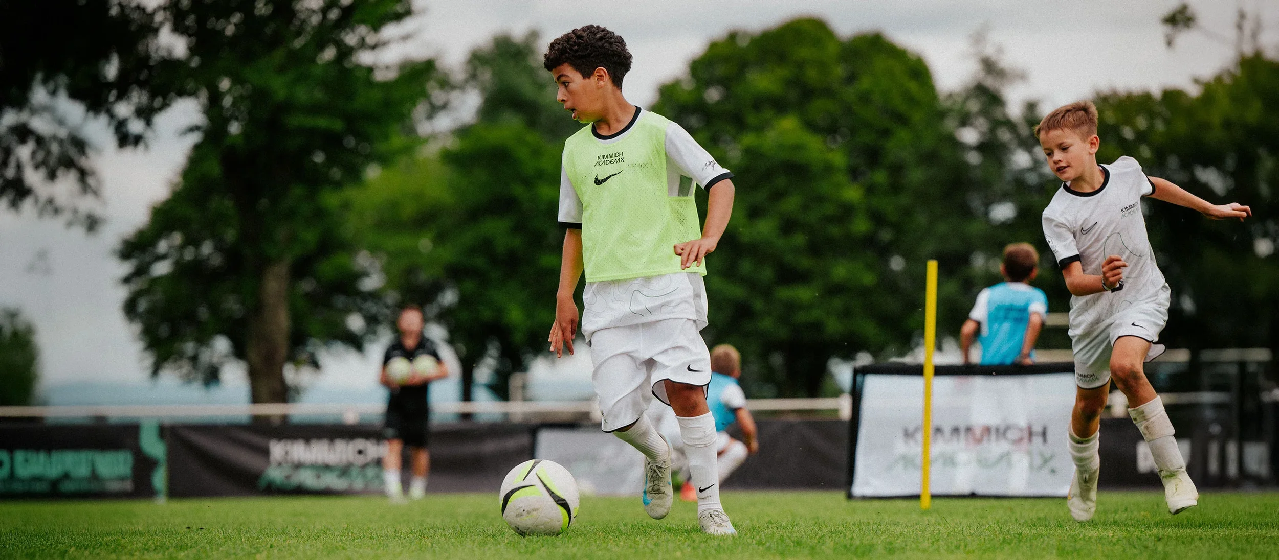 Young boys playing soccer on a field, with some running and others watching, surrounded by trees.