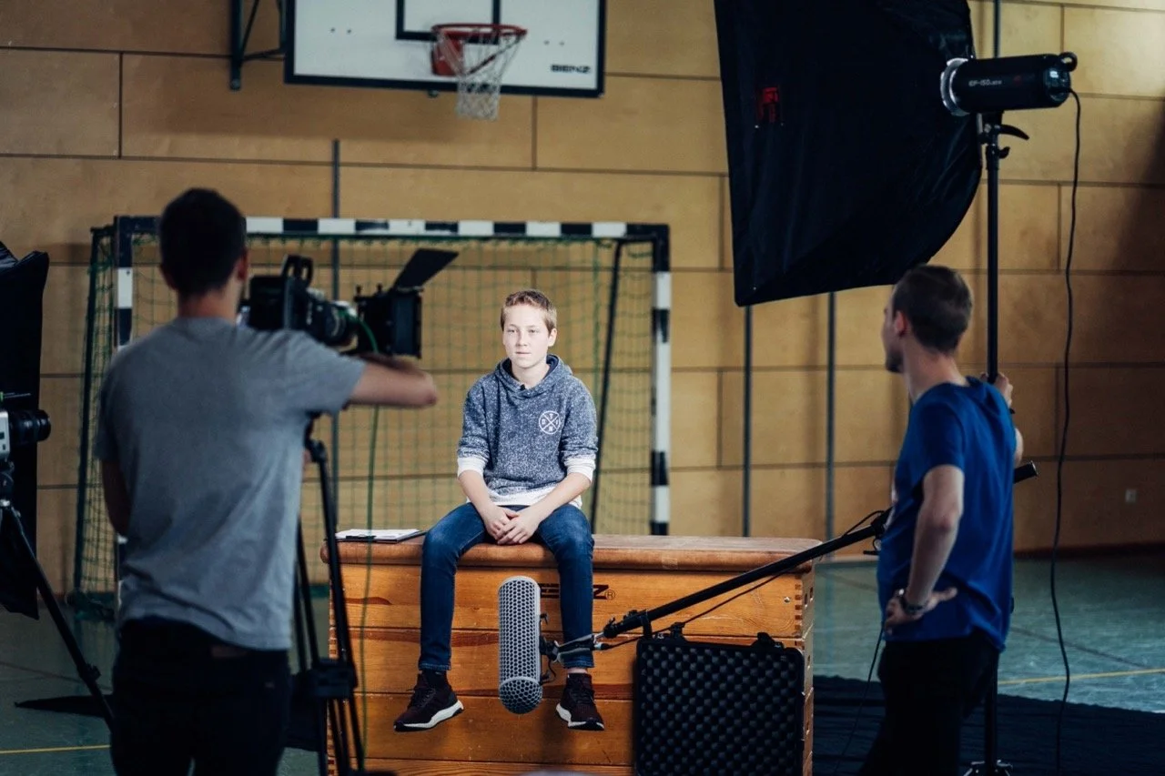 Young boy sitting on a wooden gymnast vault in a gymnasium, being filmed for a video shoot with two crew members and professional lighting.