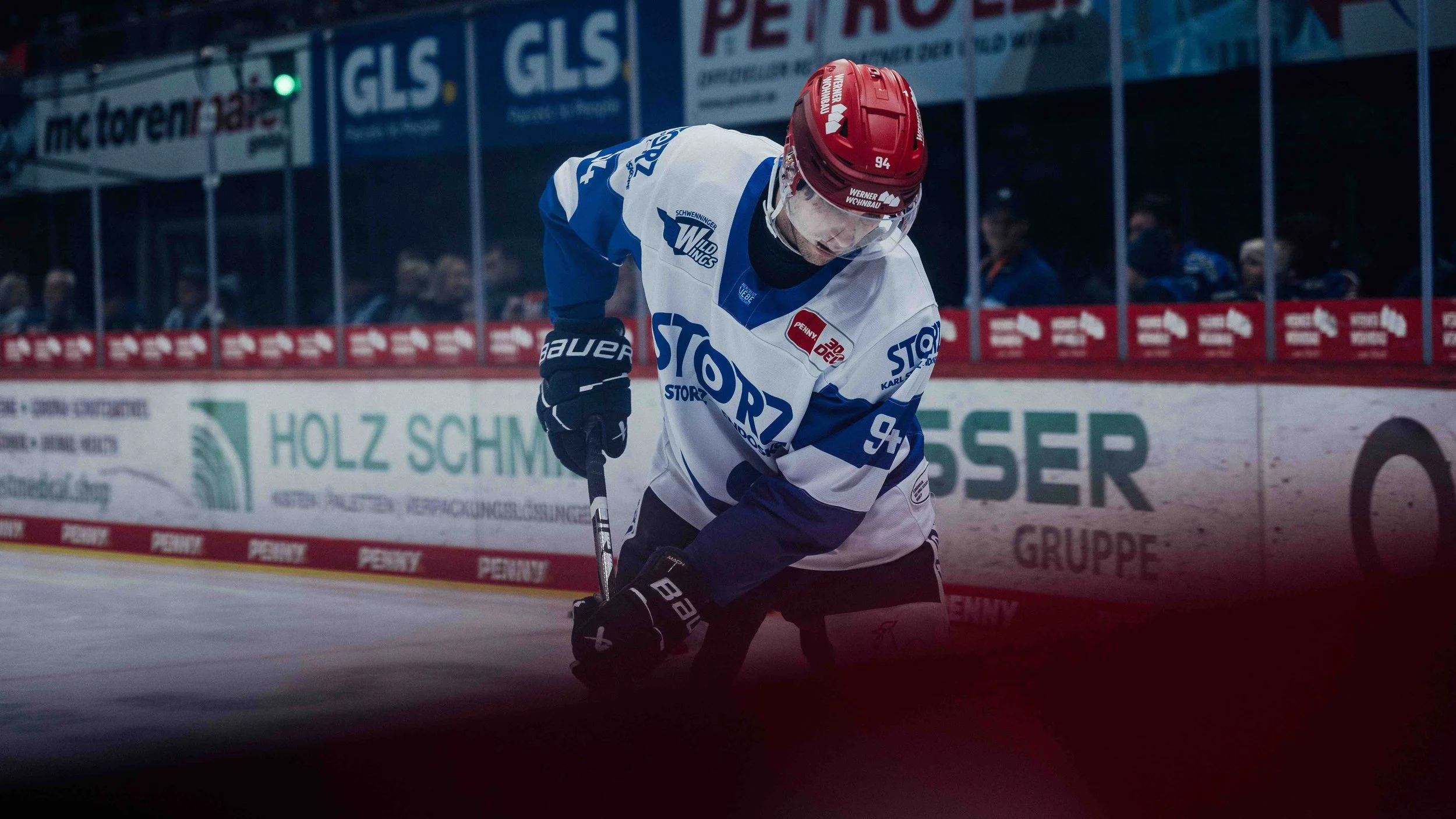 An ice hockey player in a white and blue jersey kneels on the ice, holding a hockey stick, with spectators watching from behind the rink glass.