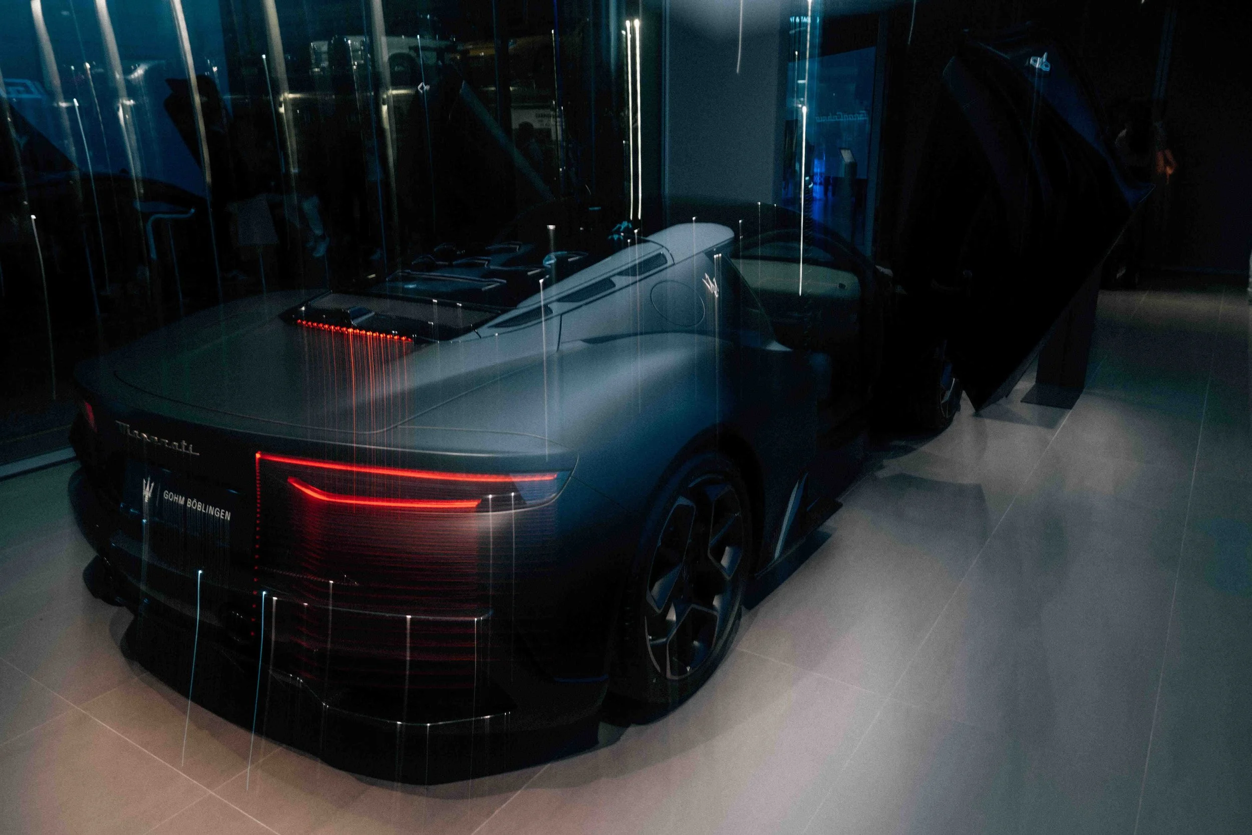A sleek black luxury sports car displayed indoors with reflective glass panels behind it.