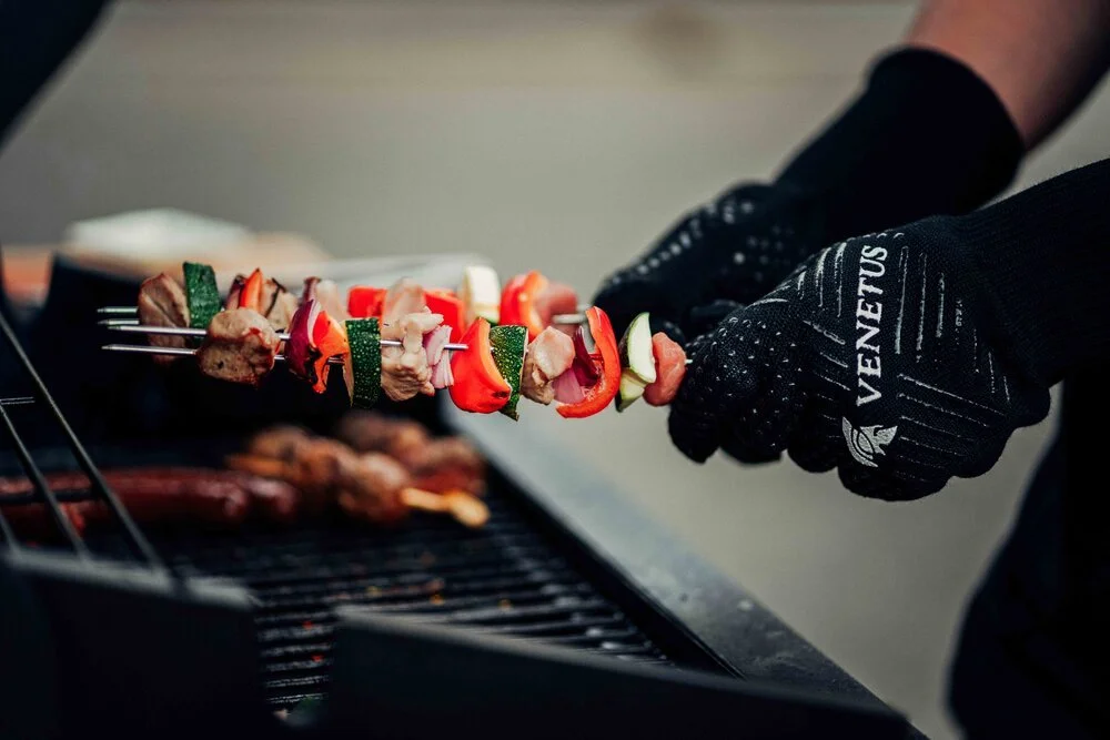 Person wearing black gloves grilling meat and vegetable skewers on a barbecue grill.