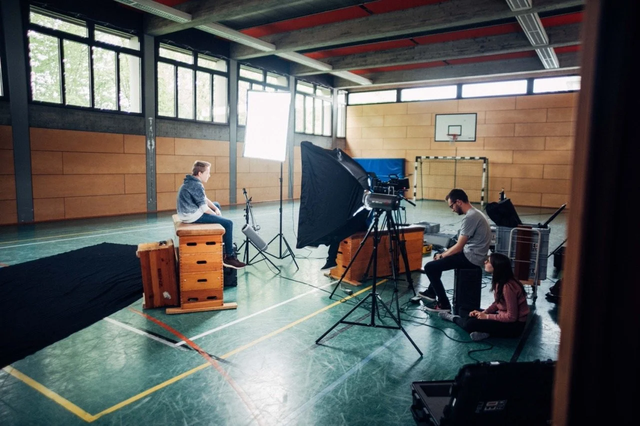 A video shoot setup with a young girl being filmed in a gymnasium, featuring lighting and camera equipment, with a man and girl working on the technical setup.