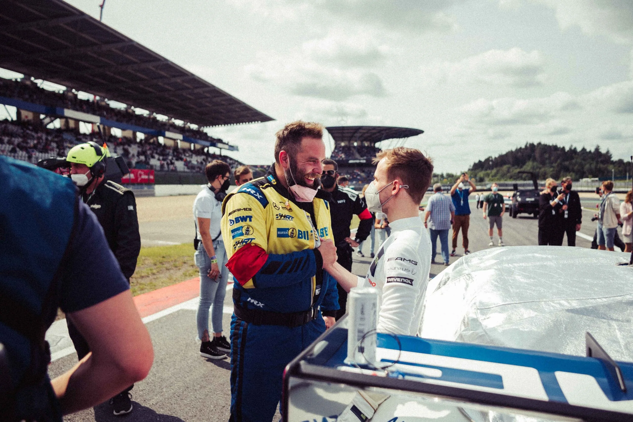 Two race car drivers, one in blue and yellow racing suit and the other in white, greet each other in the pit lane at a race track, with spectators and team members in the background. Both are wearing face masks.