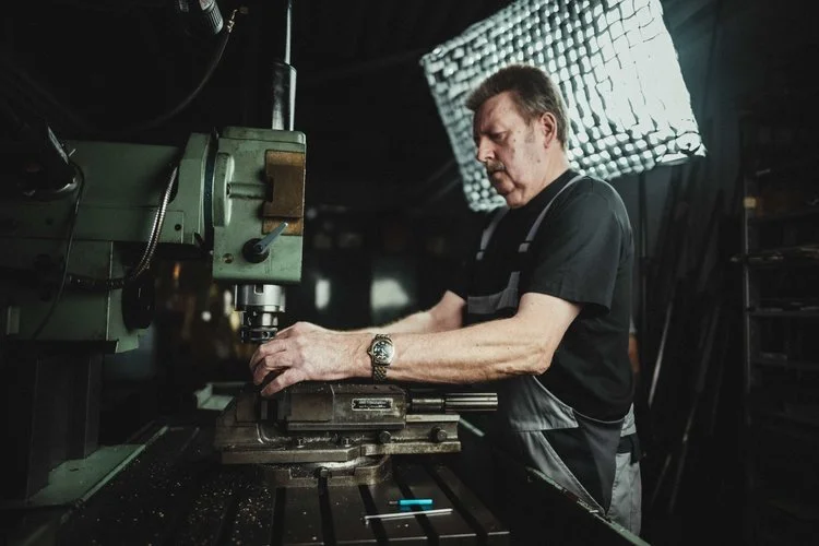 A man working with a milling machine in a workshop, wearing a black shirt and apron, with a bright light source behind him.