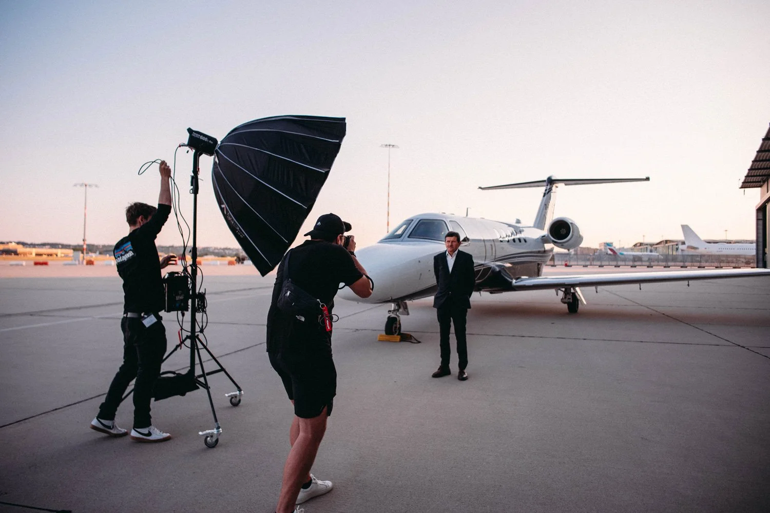 A man in a suit is standing in front of a private jet on an airport tarmac during sunset, while two crew members are preparing photography equipment, including a large umbrella reflector, for a photo shoot.