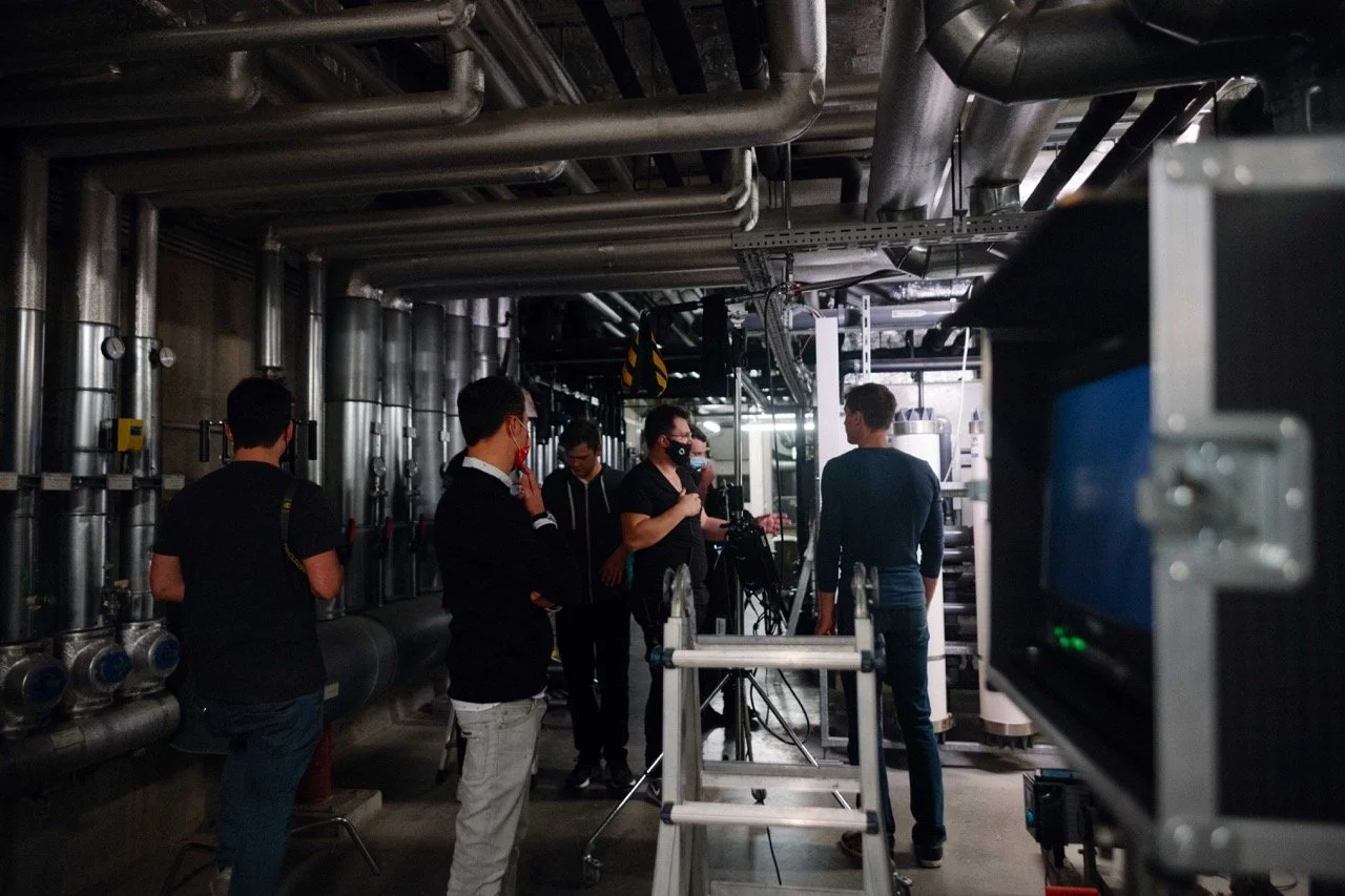 Group of people in an industrial setting with pipes, engaged in a filming or photography session. Some are wearing masks, operating cameras and equipment.