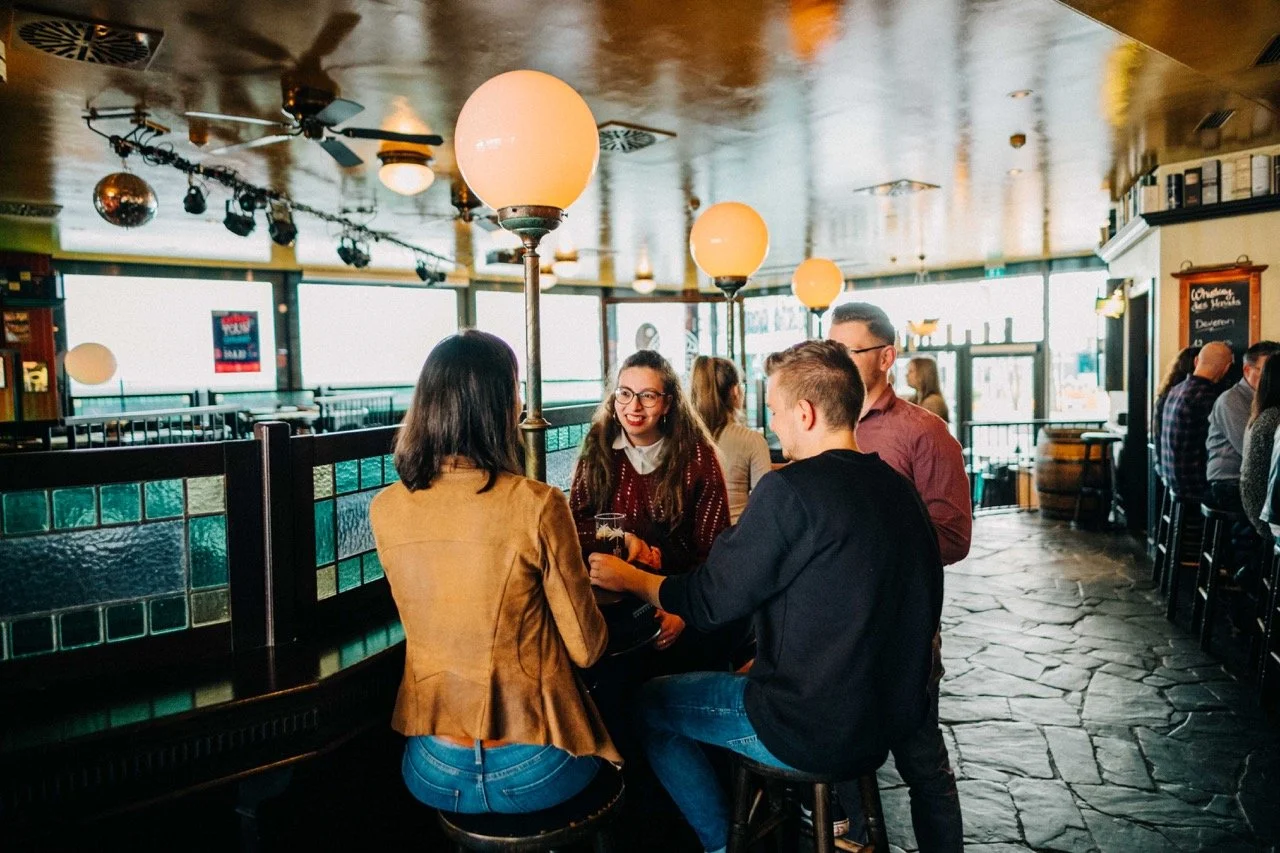 Group of four friends talking and laughing around a bar table in a lively pub, with other patrons seated along the bar behind them.