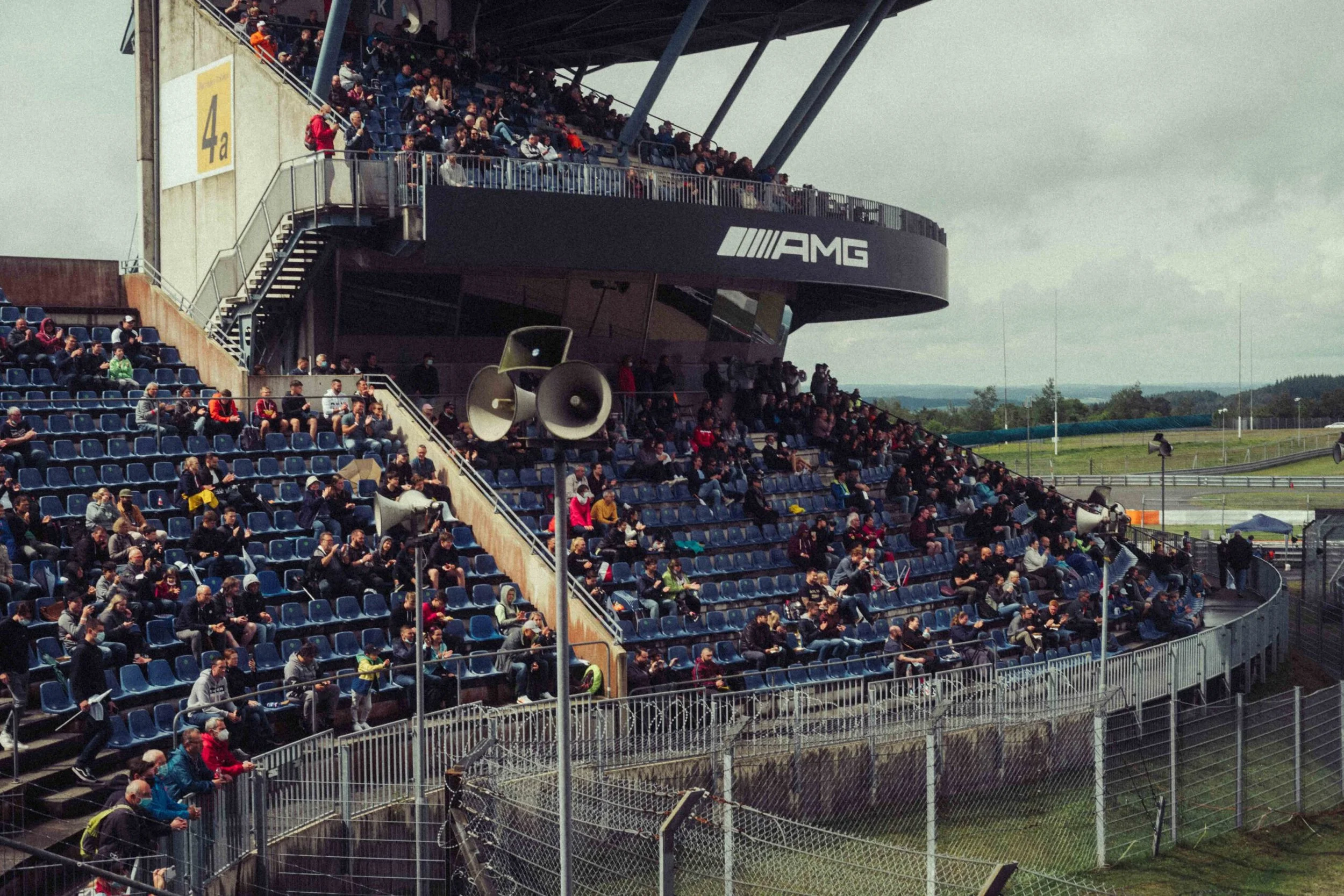 Crowd seated in the grandstand at a racing circuit on a cloudy day, with speakers and an AMG logo visible.