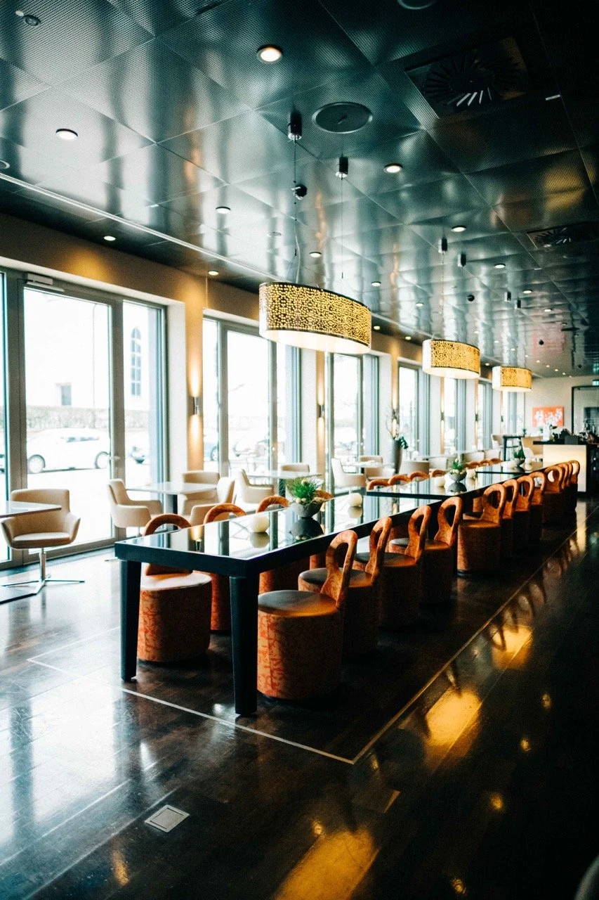 Interior of a modern restaurant or cafe with large windows, hanging pendant lights, and a long black table surrounded by upholstered chairs.