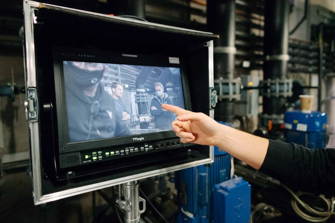 Person pointing at a monitor displaying a scene with three people in an industrial setting.