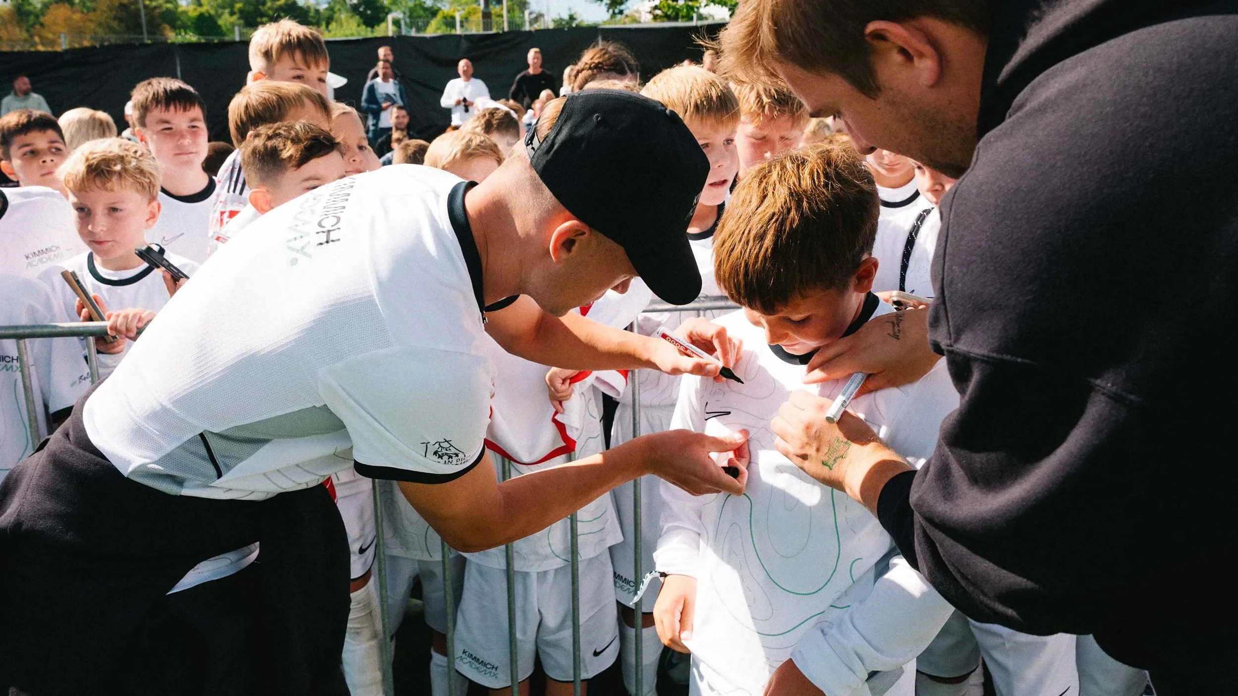 A group of young boys and two adult men, likely coaches or event organizers, gather around a boy who is signing autographs on his shirt at an outdoor sports event, with other children and spectators in the background.