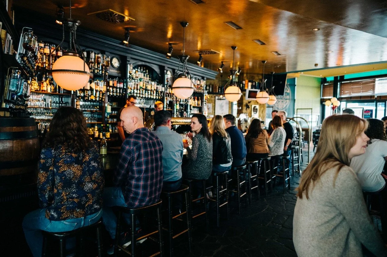 People sitting at a bar inside a restaurant, with shelves of alcohol bottles behind the bar and hanging pendant lights overhead.