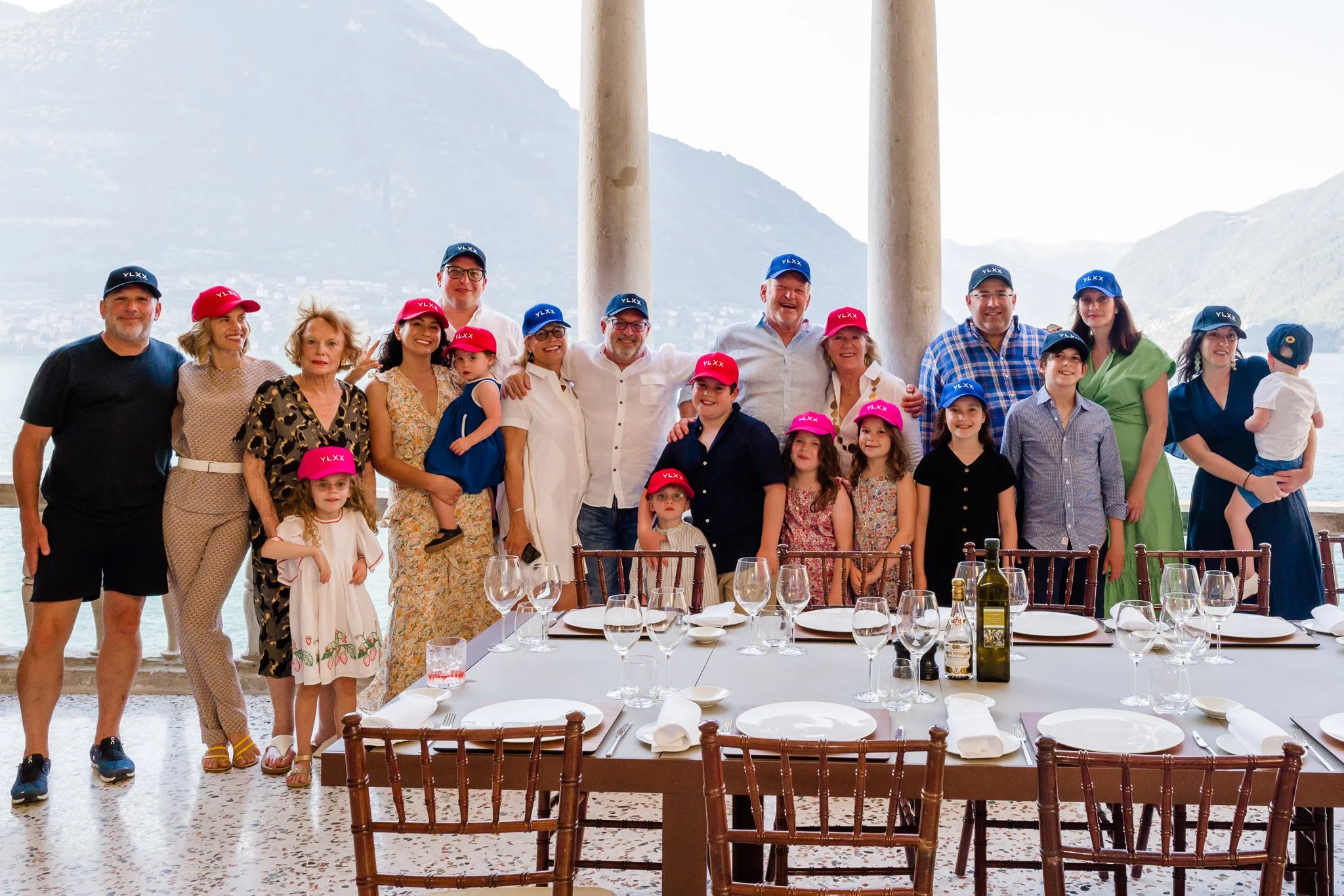 Family and friends posing for a group photo at a celebration, wearing colorful hats with initials, seated at a beautifully set dining table with wine glasses and bottles, overlooking a mountain and lake background.