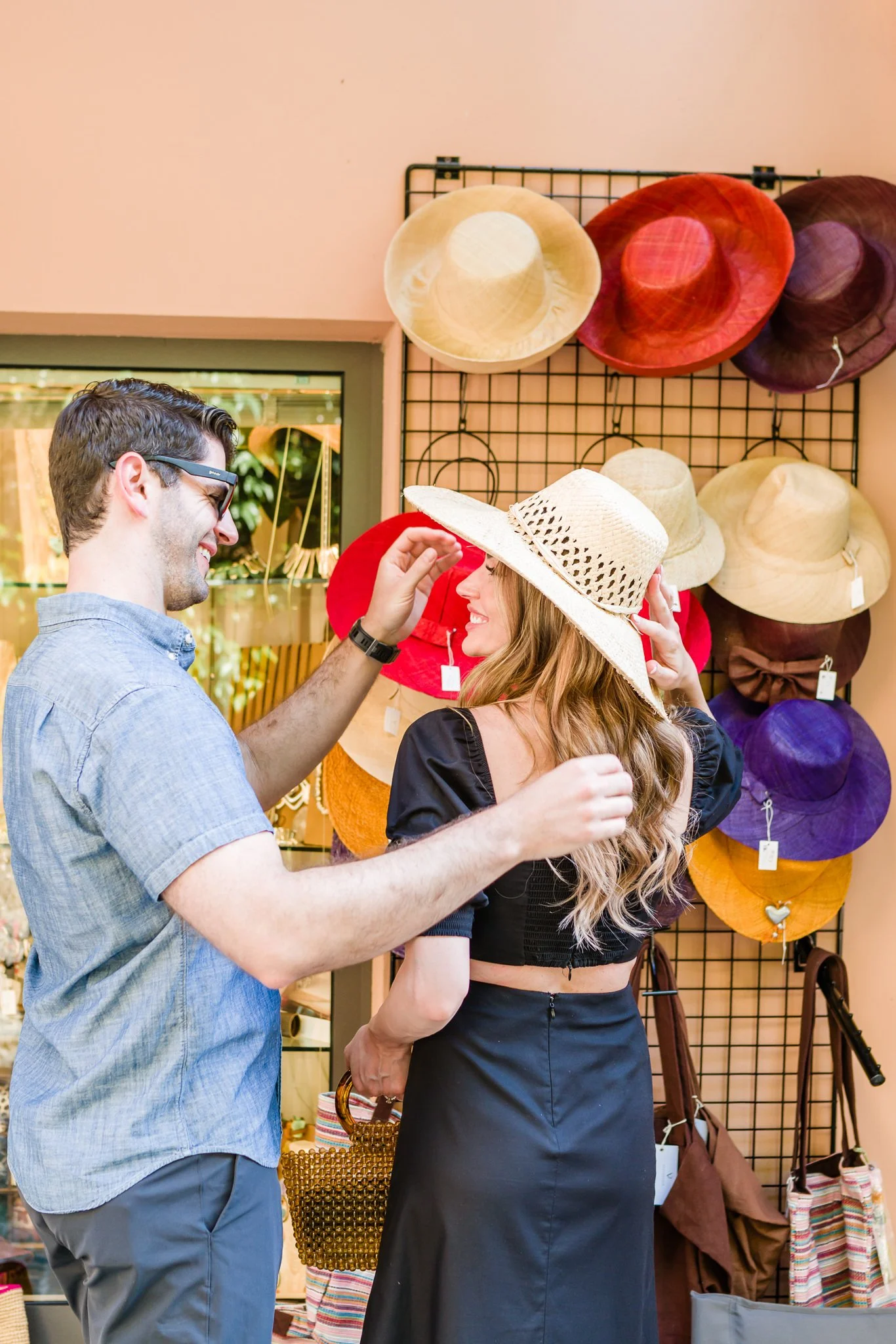 A man and woman shopping for hats in a store, with the woman trying on a large straw sunhat.