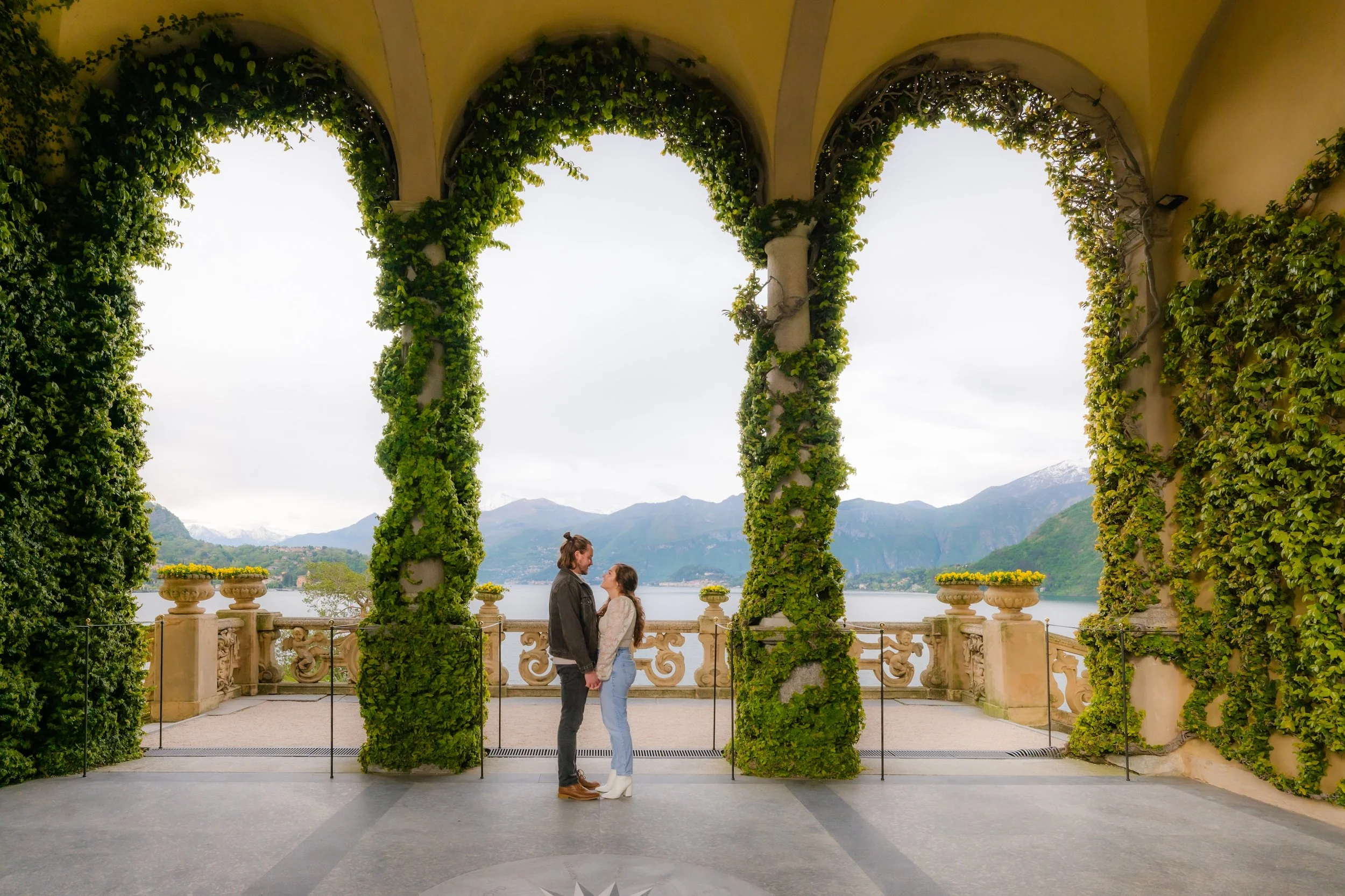 A couple standing close together and holding hands on a balcony with a scenic view of mountains and a lake, framed by arches covered in greenery.