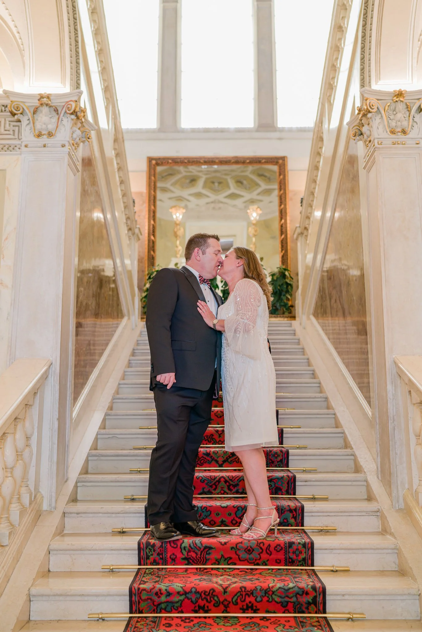 A couple dressed in wedding attire sharing a kiss on a staircase inside a grand, elegant building with ornate decor and large windows.