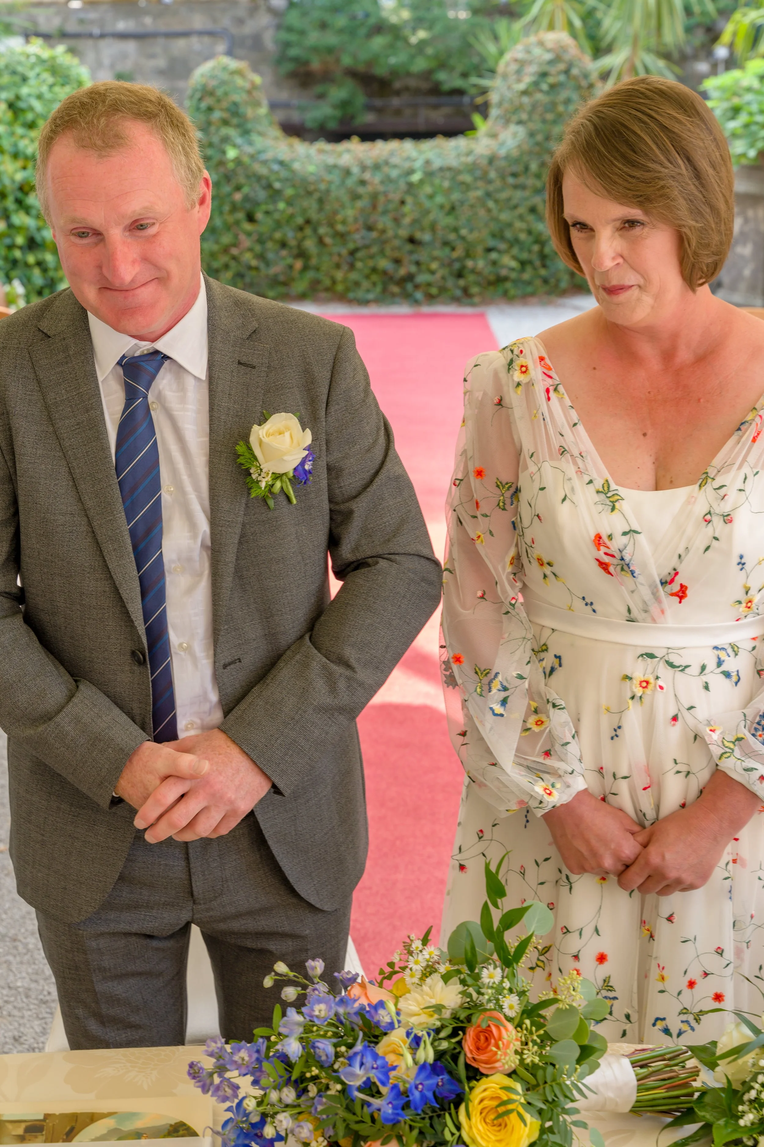 A man and woman standing at a wedding ceremony, the man is wearing a grey suit with a white shirt, patterned blue tie, and a white rose boutonniere. The woman is wearing a white dress with colorful floral embroidery. A table with a bouquet of flowers