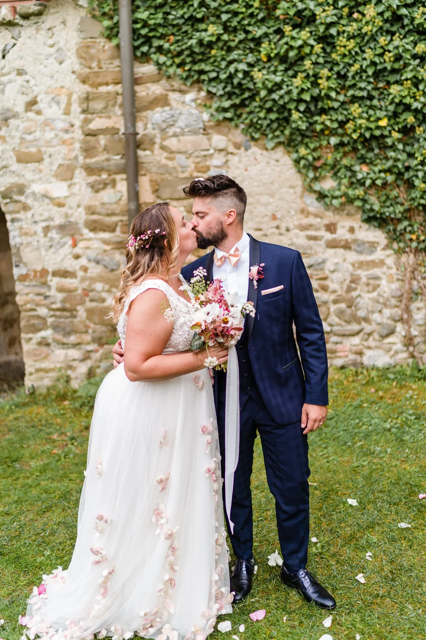 A bride and groom sharing a kiss outdoors during their wedding, with a stone wall and greenery in the background. The bride holds a bouquet of flowers, and the groom wears a dark blue suit with a light-colored bow tie.