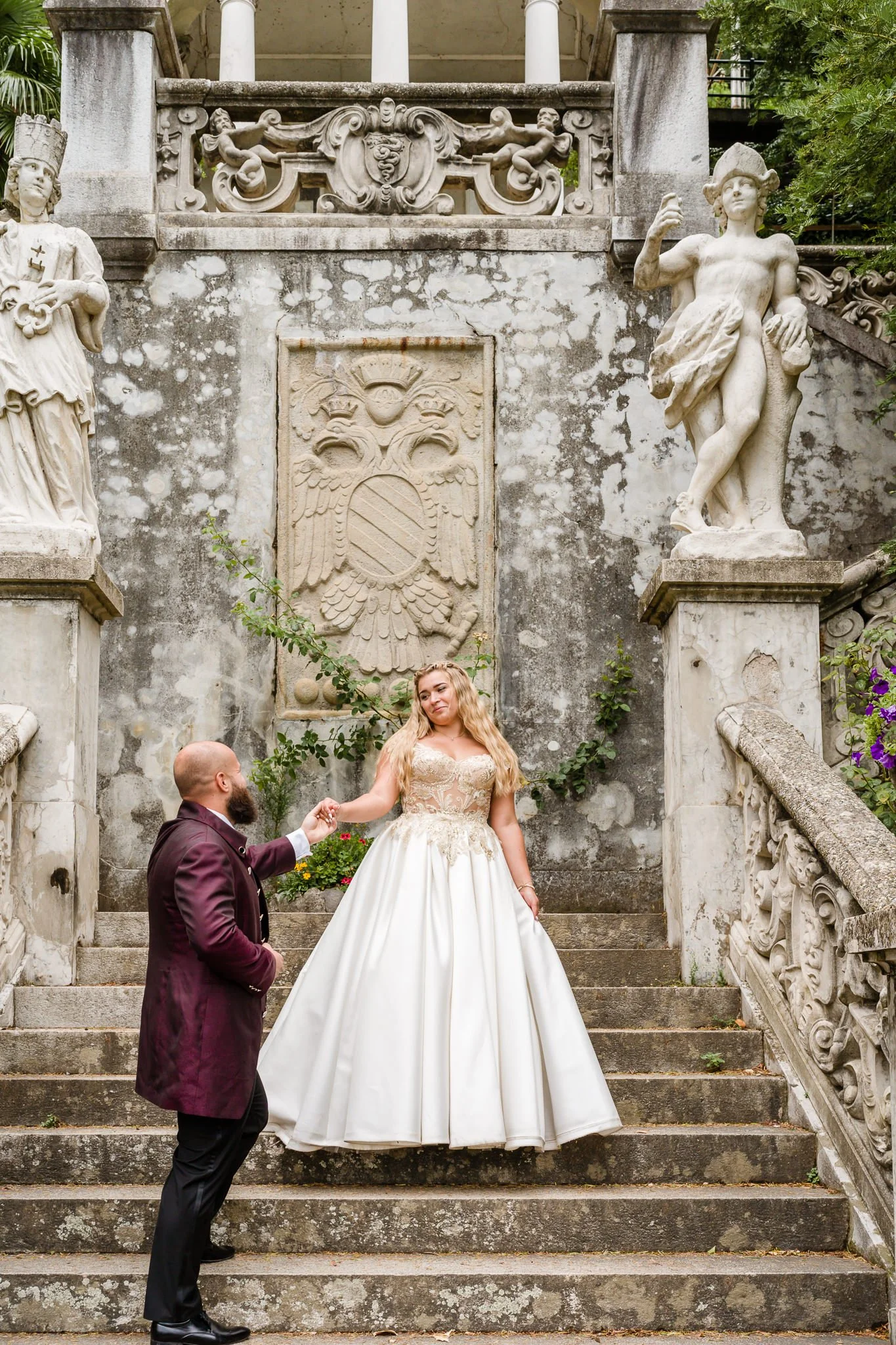 A couple, with the man kneeling and the woman standing in a wedding dress, is on the steps of a historic stone staircase with classical statues and sculptures surrounding them.