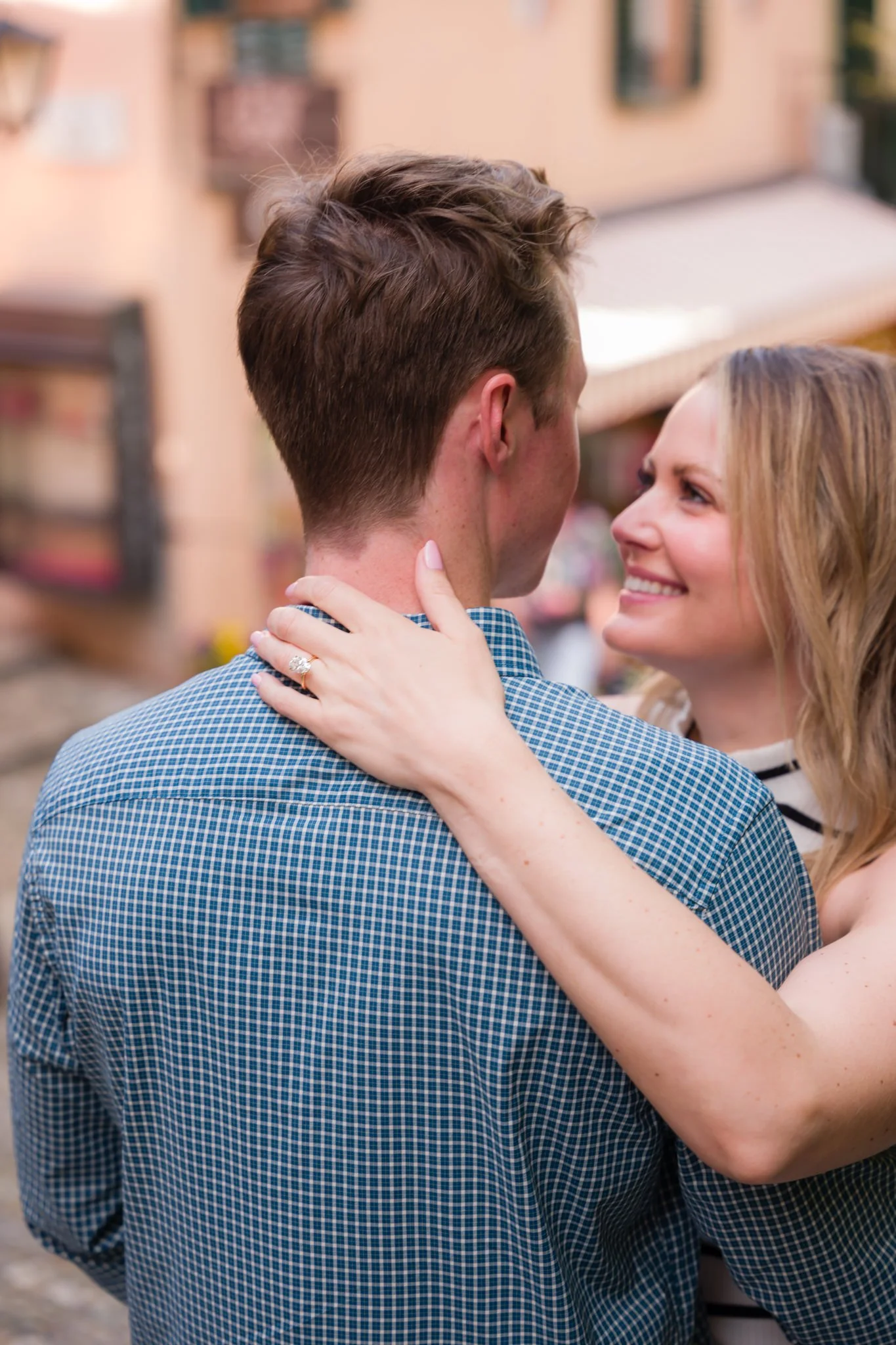 A woman with blonde hair smiling while holding a man with short brown hair, outdoors in a town.