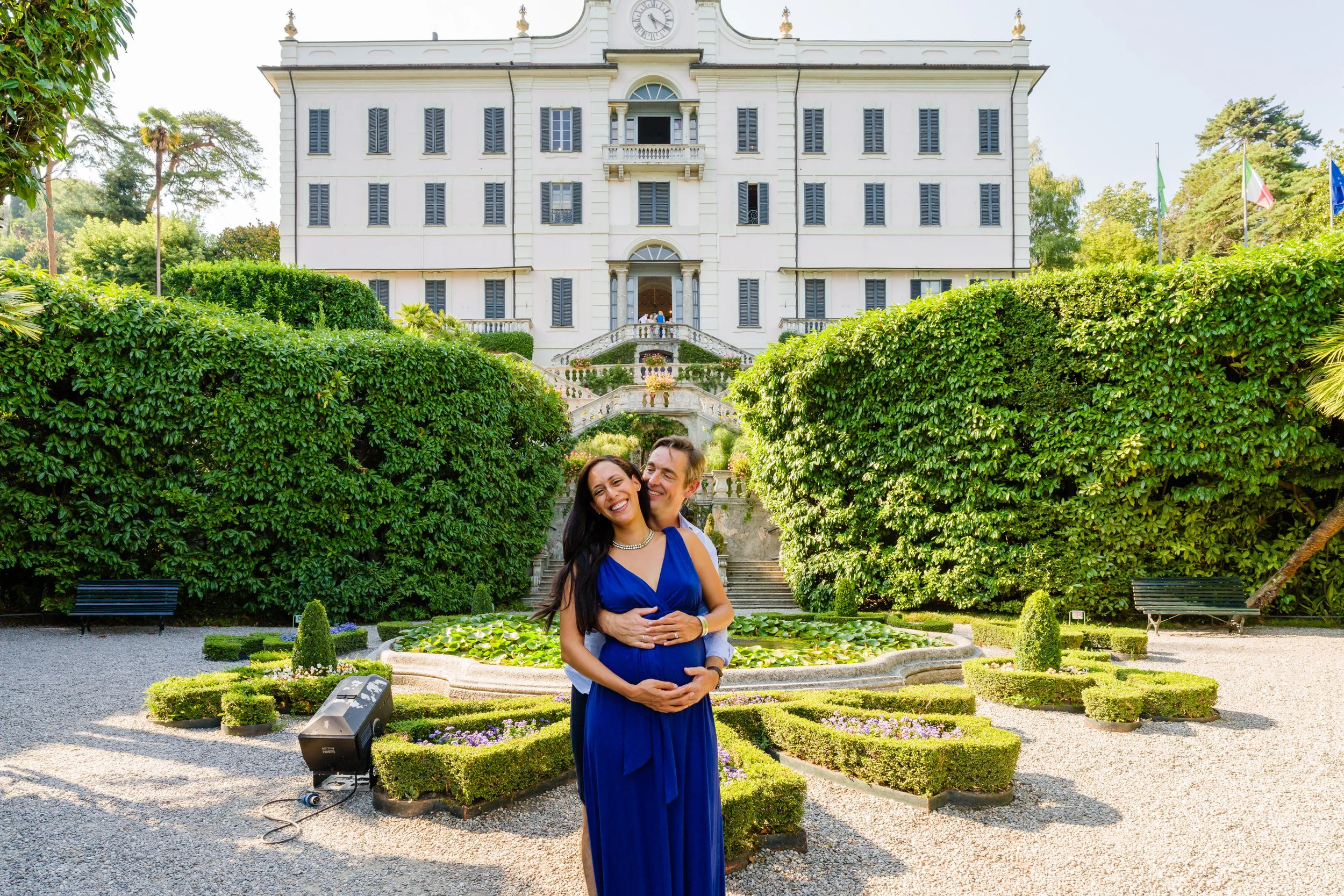 A couple, an African American woman and a man, standing in a garden with a large white building in the background, smiling and embracing, the woman pregnant, dressed in blue. The garden has neatly trimmed bushes, a pond, and gravel paths.