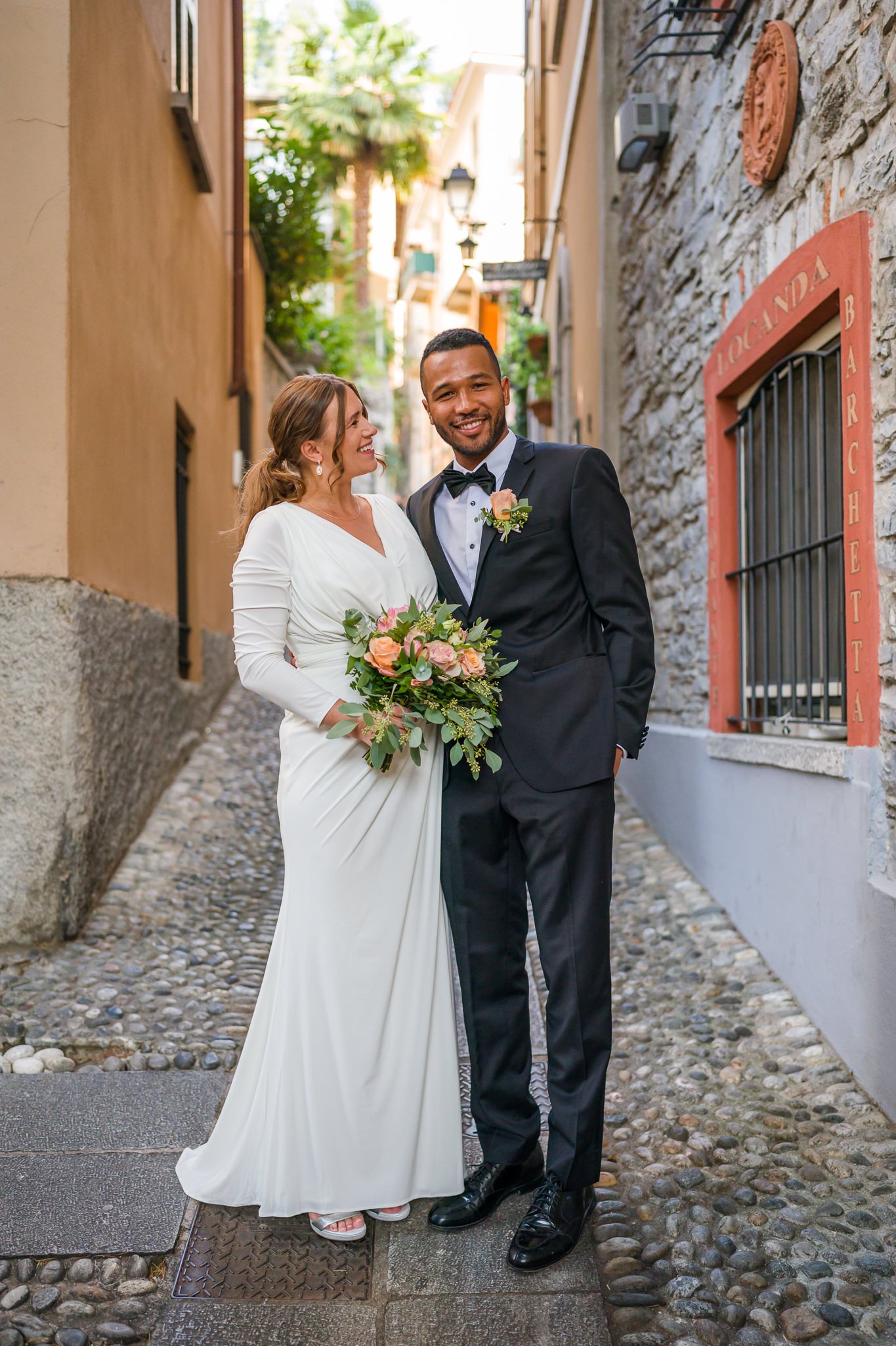 A newlywed couple standing on a cobblestone street in a charming European alley, smiling and looking at each other. The bride wears a white wedding dress and holds a bouquet of pink and peach flowers, while the groom wears a tuxedo with a black bow t