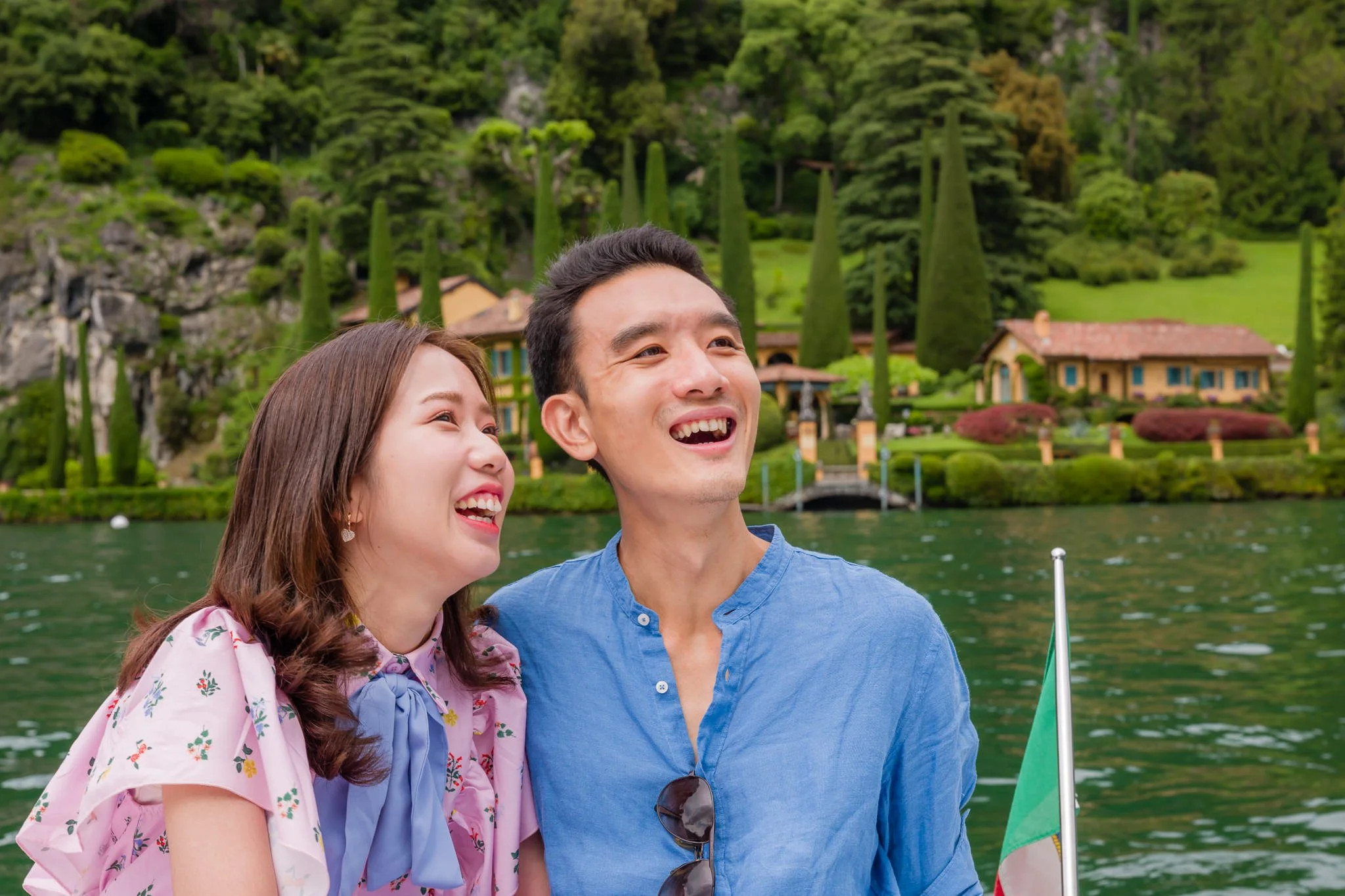A happy couple smiling on a boat with a scenic lakeside mansion and lush green trees in the background.