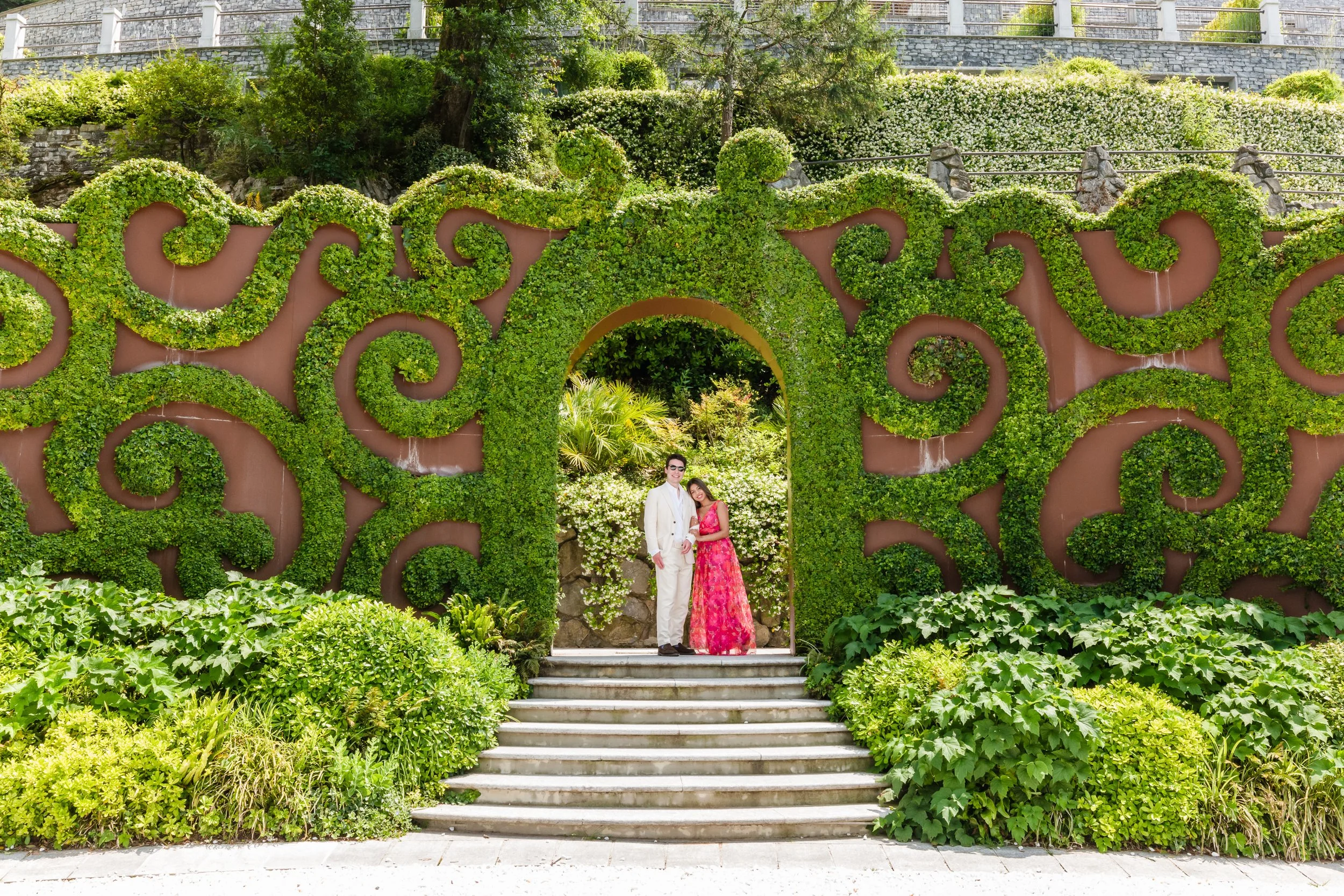 A couple dressed in formal attire posing under a decorative archway covered in green foliage with floral accents, surrounded by lush green bushes and plants, with a stone staircase leading up to the arch.