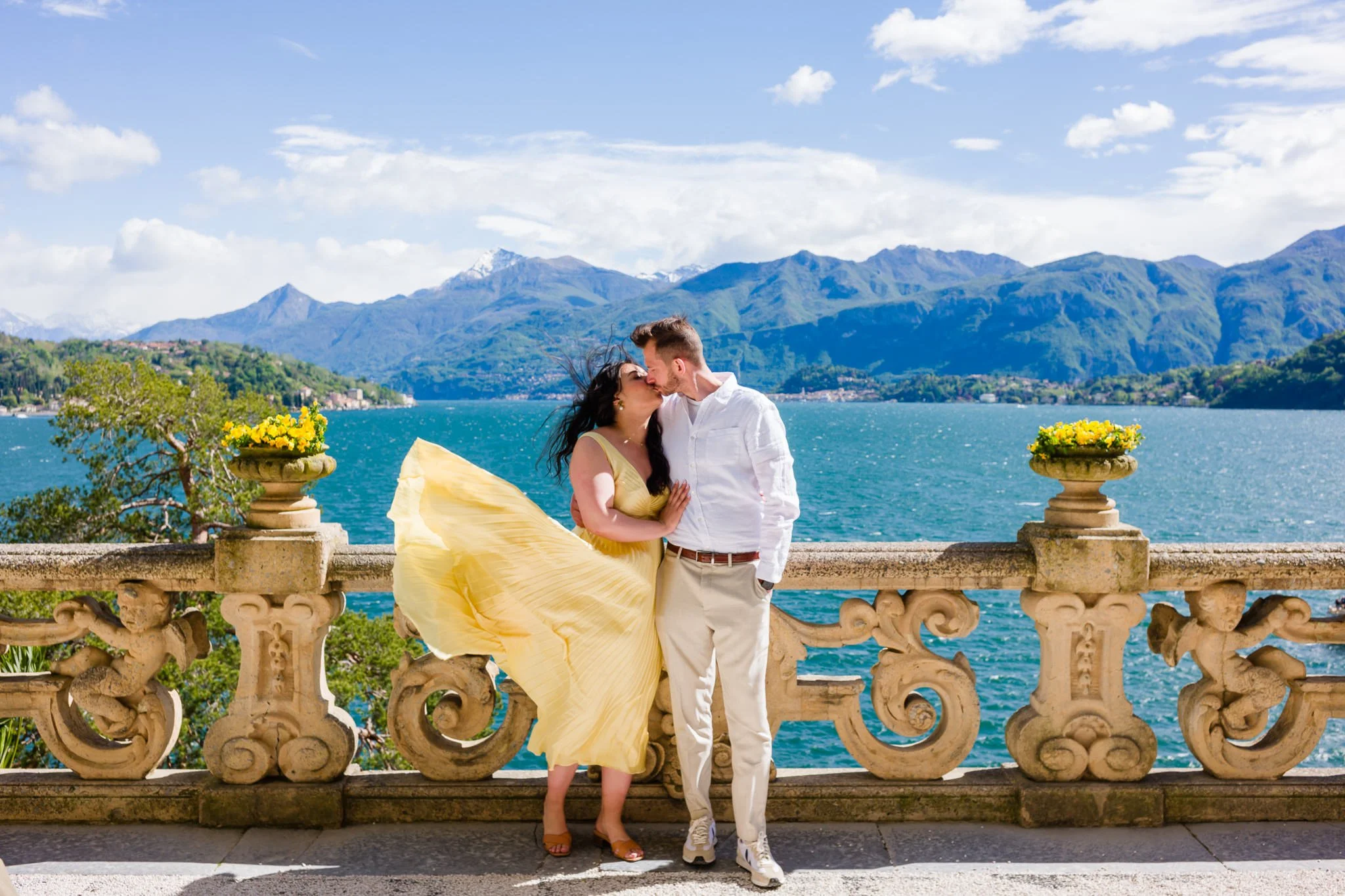 A couple stands close together on a stone balcony overlooking a lake with mountains in the background. The woman in a yellow dress is kissing the man in white. The balcony has ornate stone railings with two flower pots on either side, each filled wit