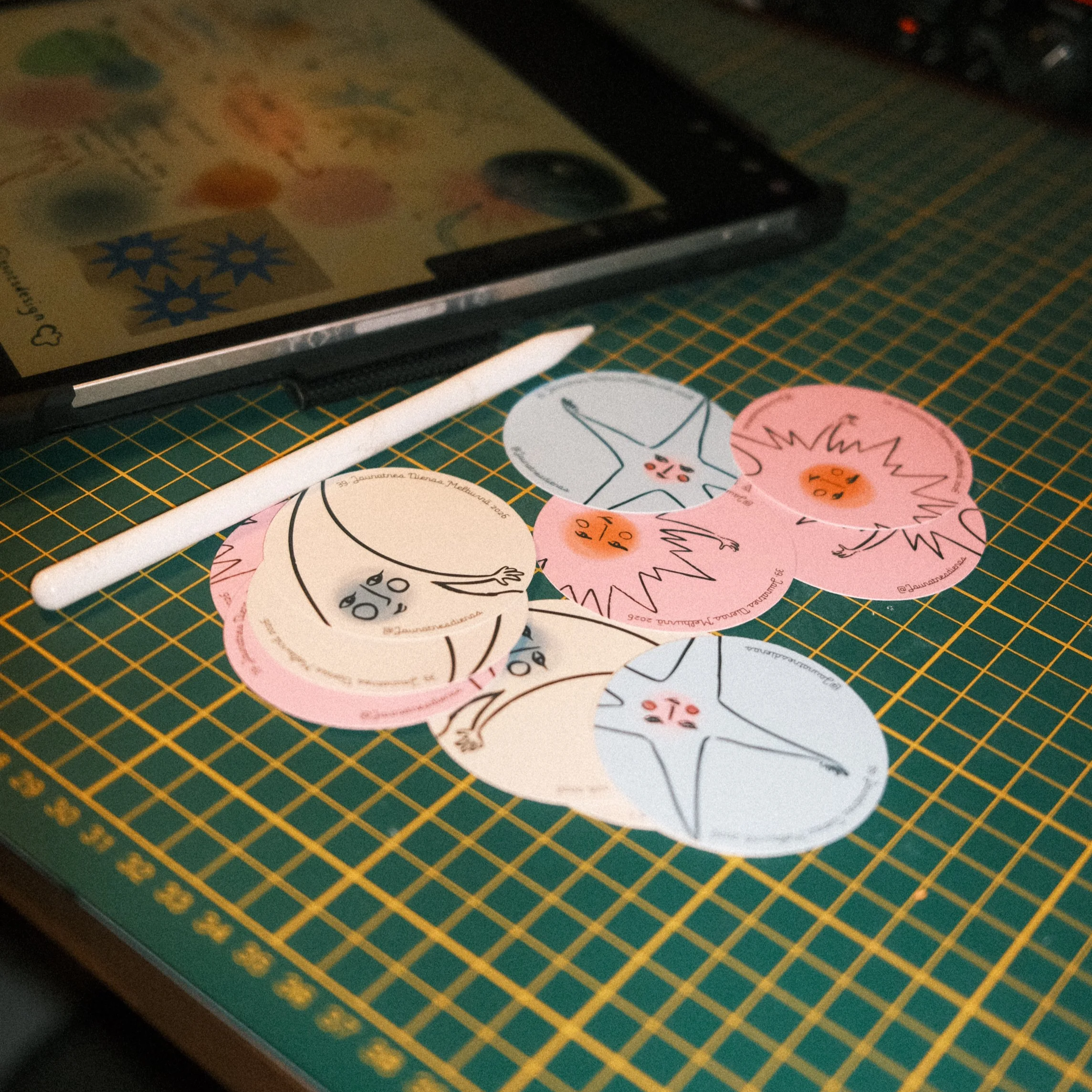 Colorful paper cutouts of starfish and sand dollars with faces, scattered on a green grid cutting mat, accompanying a white stylus and a tablet screen in the background.
