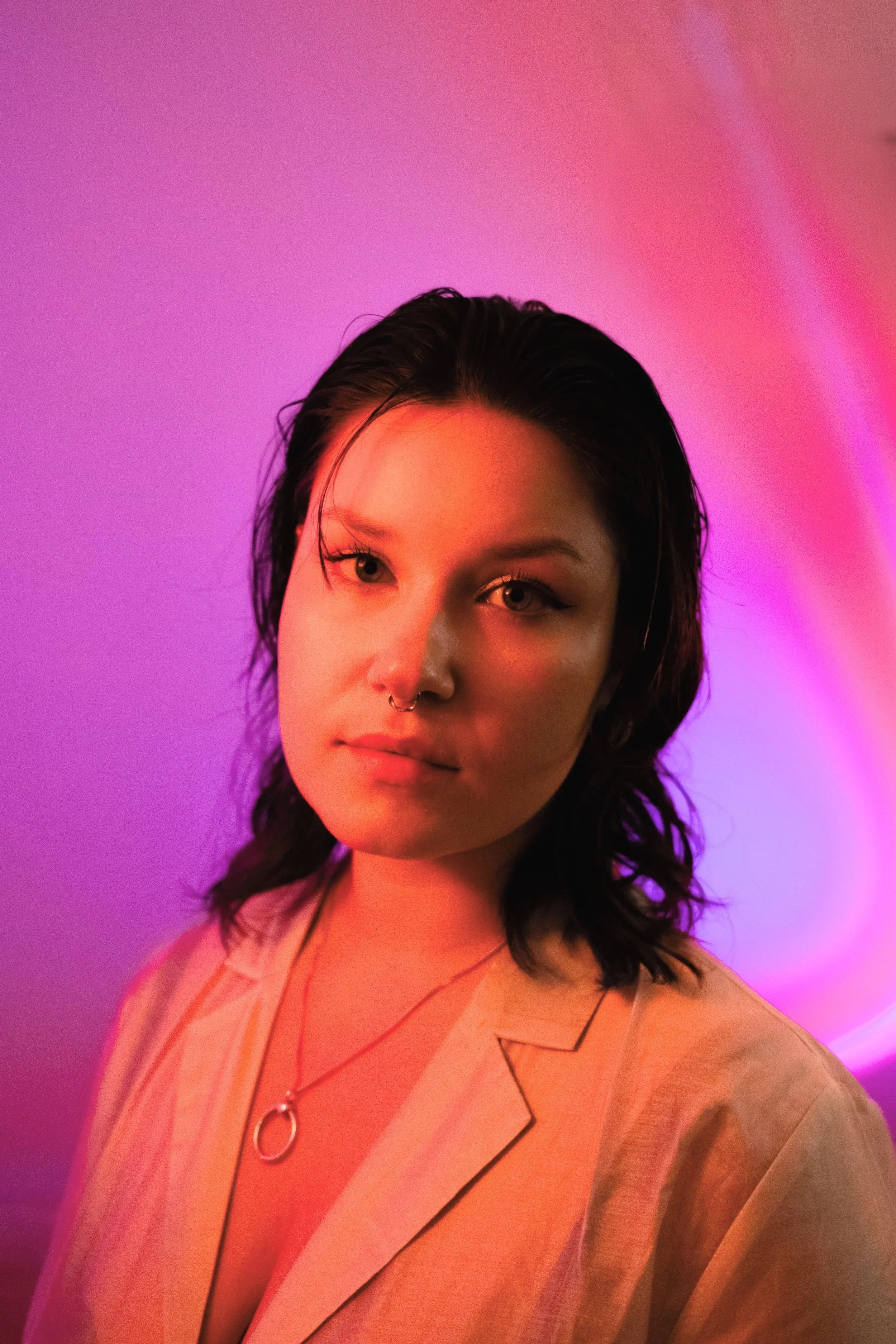 Portrait of a young woman with dark hair, wearing a beige blazer and a necklace with a circular pendant, illuminated by pink and purple neon lights.