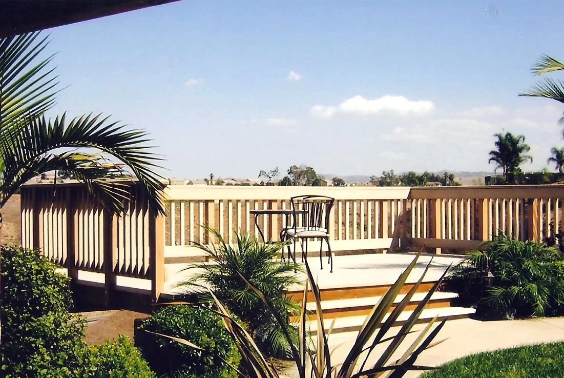 An outdoor patio with wooden railing, two chairs, and lush green plants under a blue sky with scattered clouds.