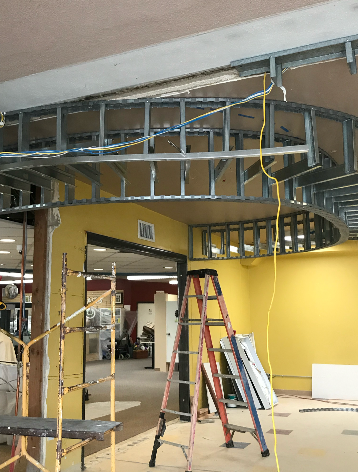 Interior of a building under construction with metal framing for a circular ceiling feature, yellow walls, and construction ladders.