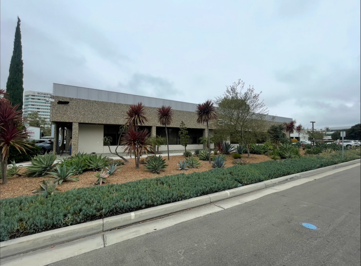 A building with a textured exterior wall, surrounded by drought-tolerant plants and bushes, on a cloudy day.
